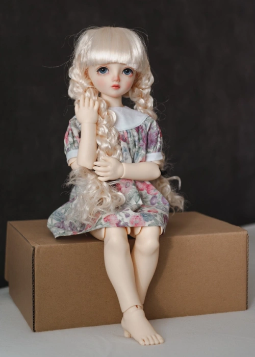 A big-eyed ball jointed doll. She has thick hair in two plaits, a gathered yoke dress, and is sitting on a cardboard box.