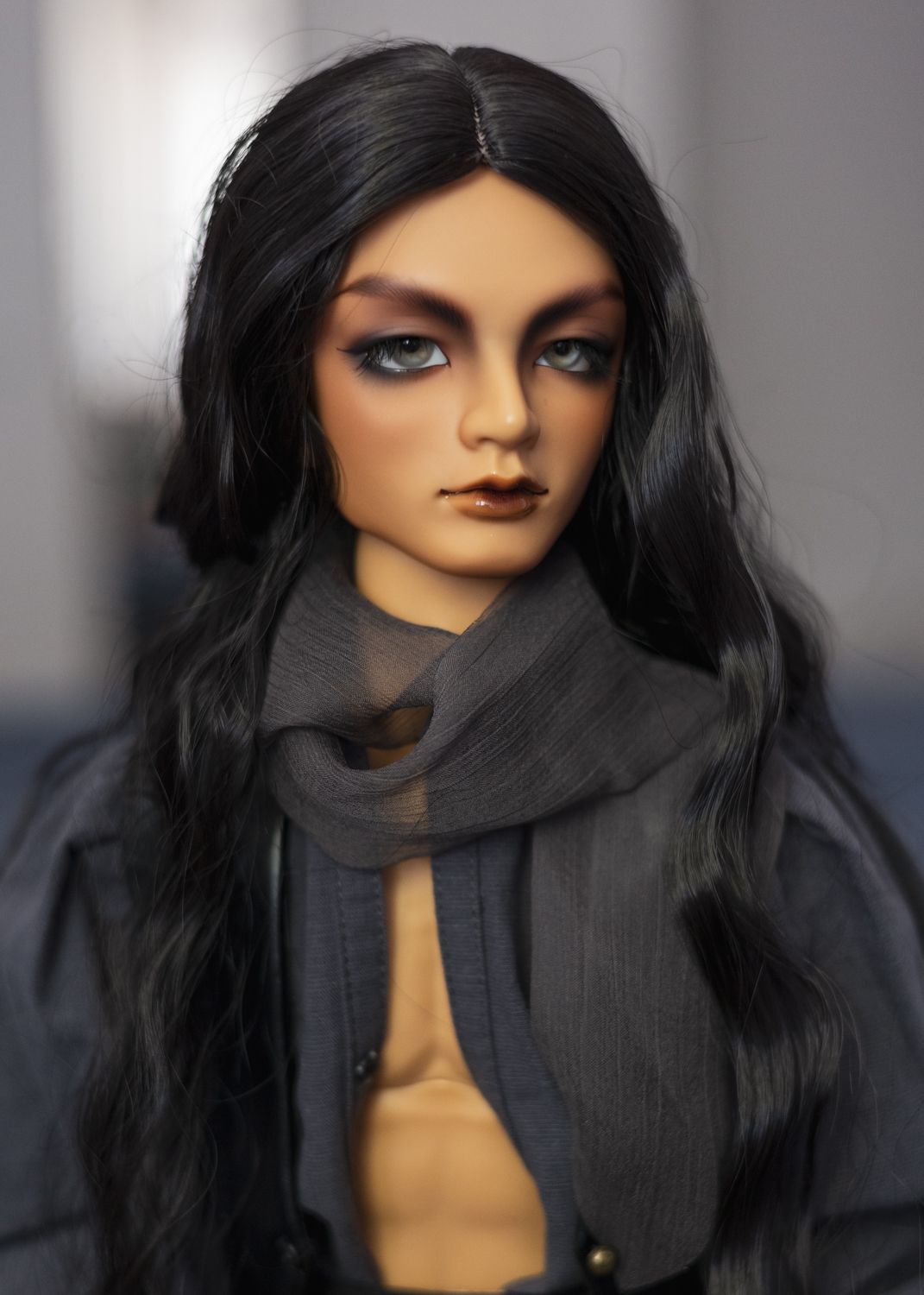 Male ball-jointed doll with tan skin and strongly-marked eyebrows, with grey eyes and long black hair. He wears an open grey shirt with a gauzy grey scarf around his neck.