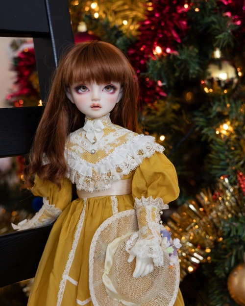 Half-length portrait of a female ball-jointed doll with long auburn hair, wearing a high-necked yellow and white dress with puffy sleeves. She leans against a human-sized chair, holds a hat in one hand, and is in front of a Christmas tree.