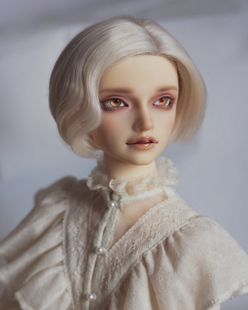 Portrait photo of a male ball-jointed doll with golden eyes, a cream chin-length wig, and a cream lacy top with ruffles and pearly buttons.