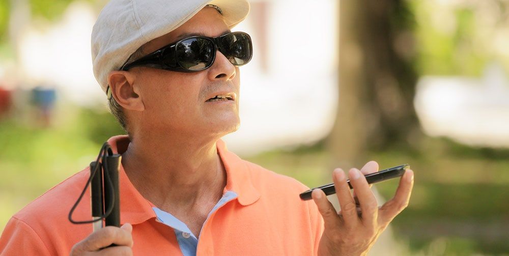 A visually impaired man wearing dark sunglasses and a white cap uses a smartphone with voice input while holding a white cane. He is outdoors, dressed in a bright orange polo shirt.