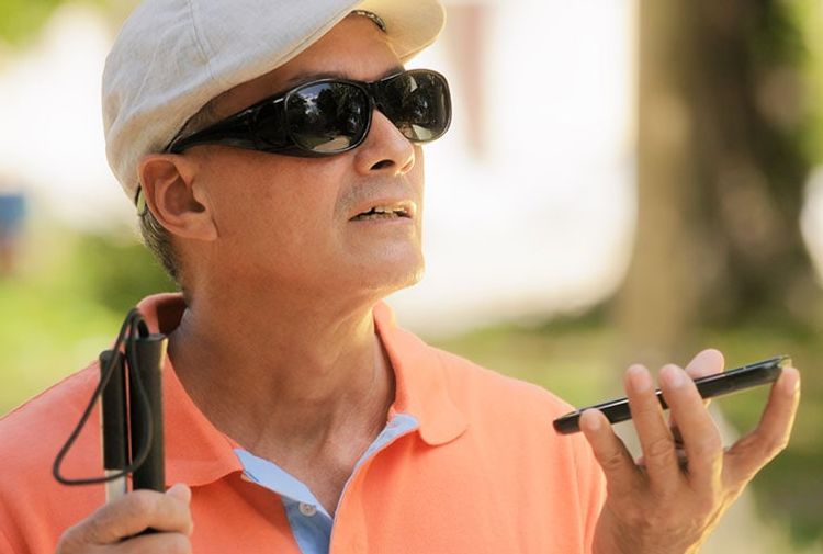 A visually impaired man wearing dark sunglasses and a white cap uses a smartphone with voice input while holding a white cane. He is outdoors, dressed in a bright orange polo shirt.