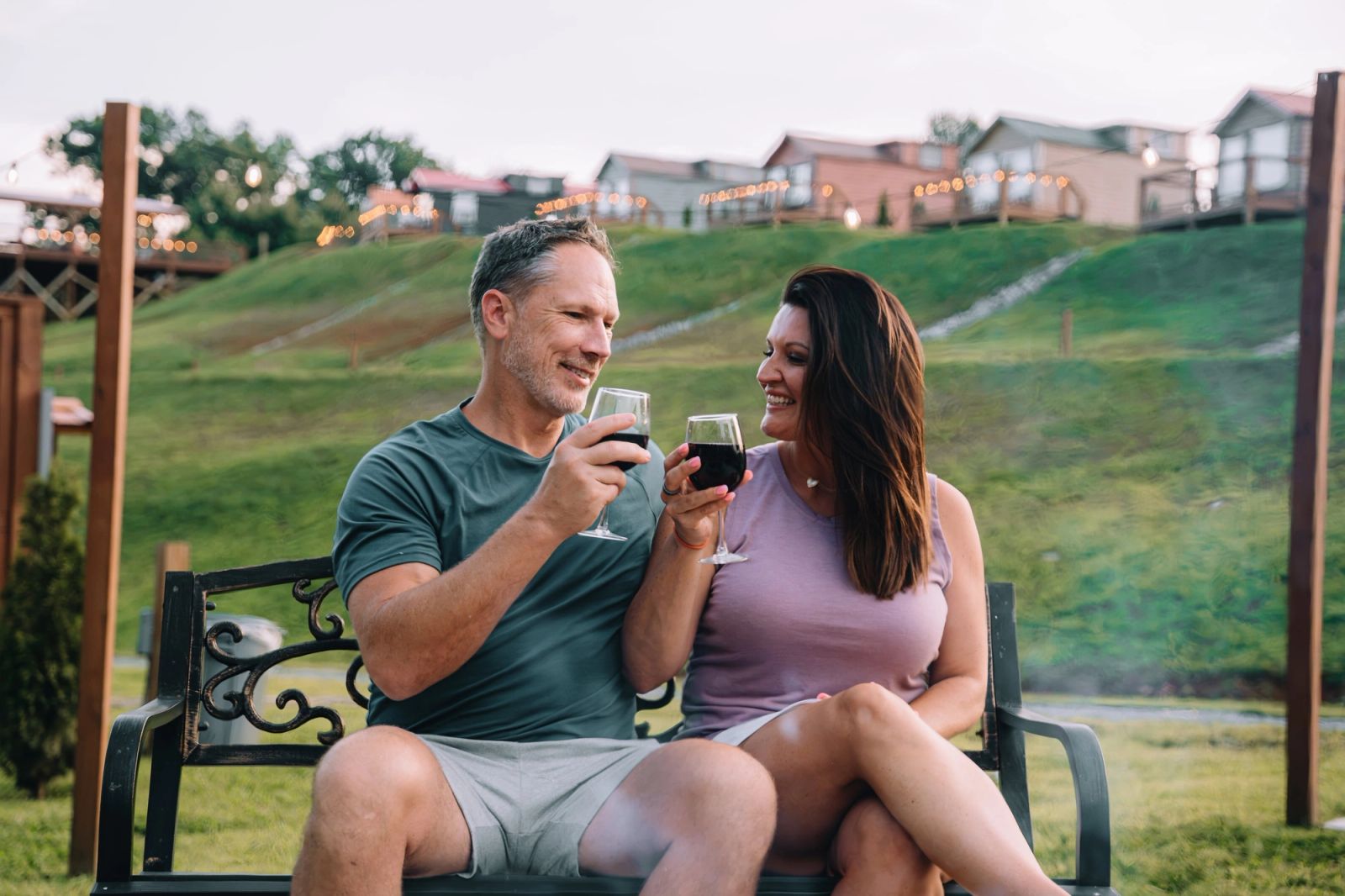 A couple toasting wine glasses beside a private firepit with tiny homes on the ridge above