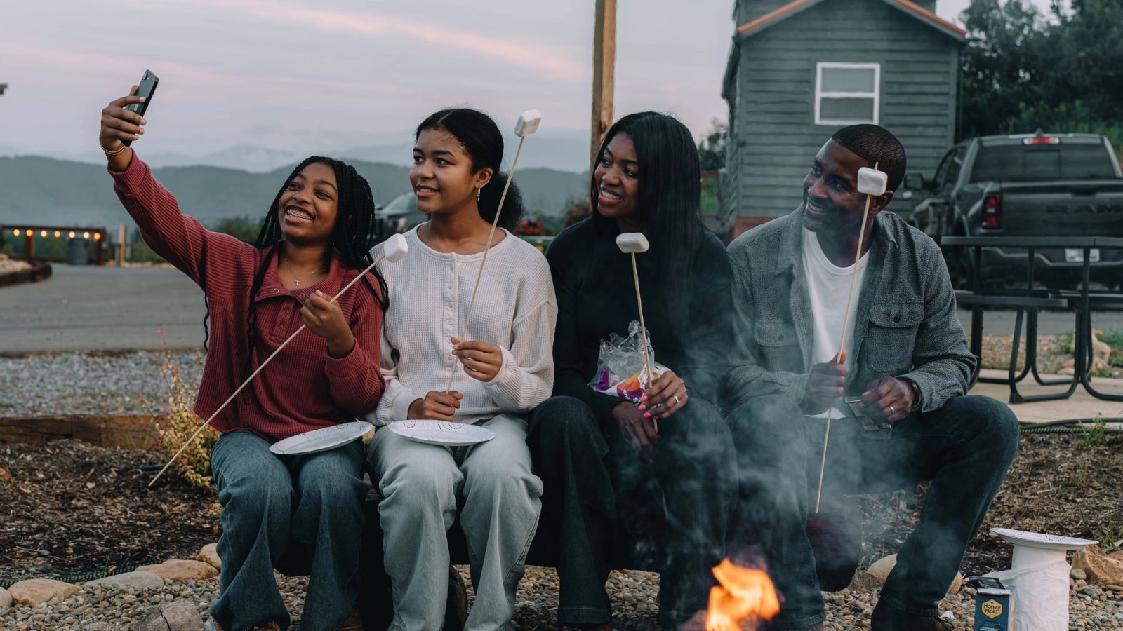 Four friends roasting marshmallows on the firepit bench, one taking a selfie, at Sojourn Lodging