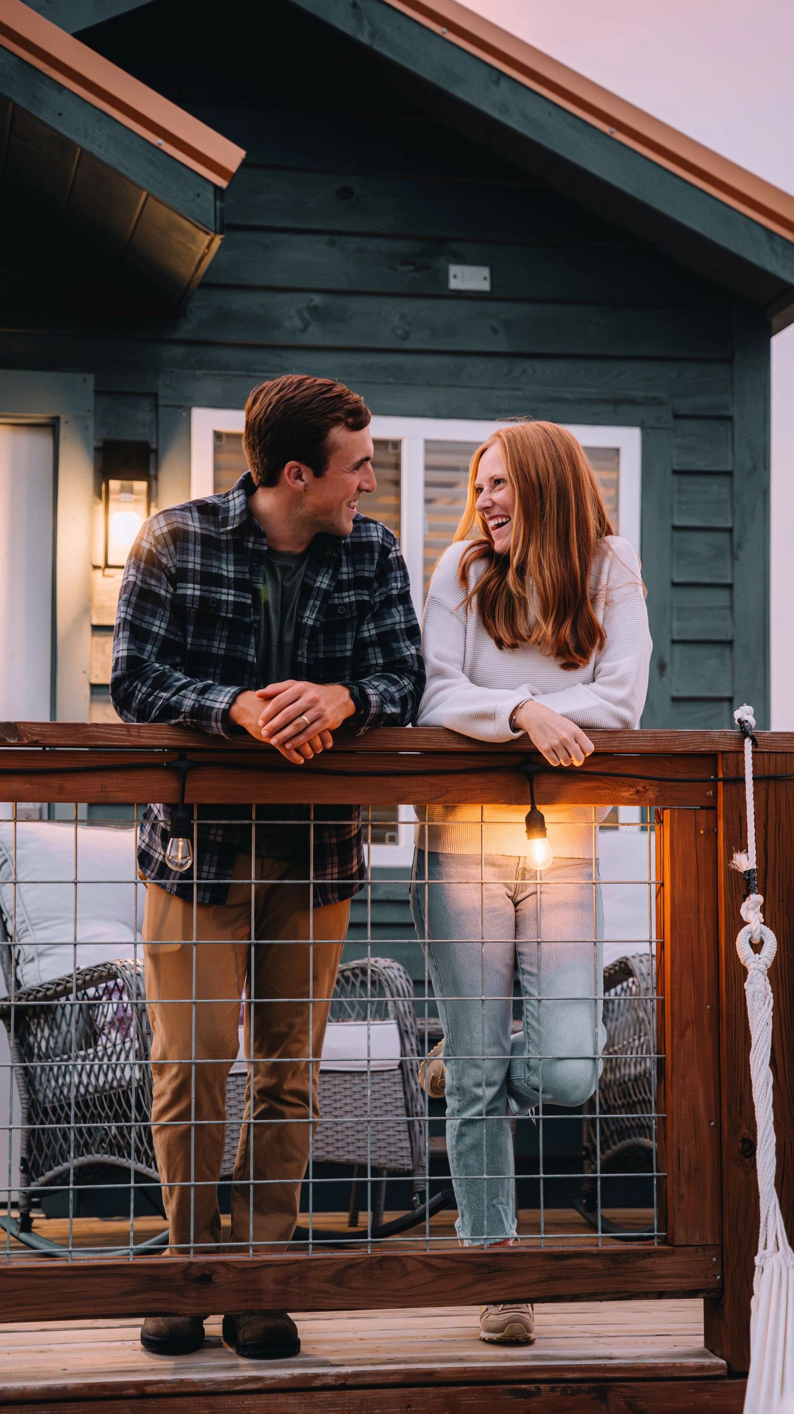 A couple talking on the string-lit deck of their tiny home at Sojourn Lodging as the sky turns pink