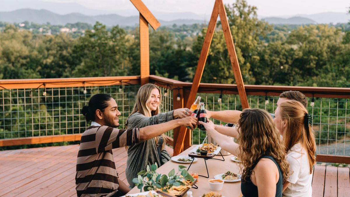 Friends toasting at the Observation Deck table beneath the timber arch, mountains beyond
