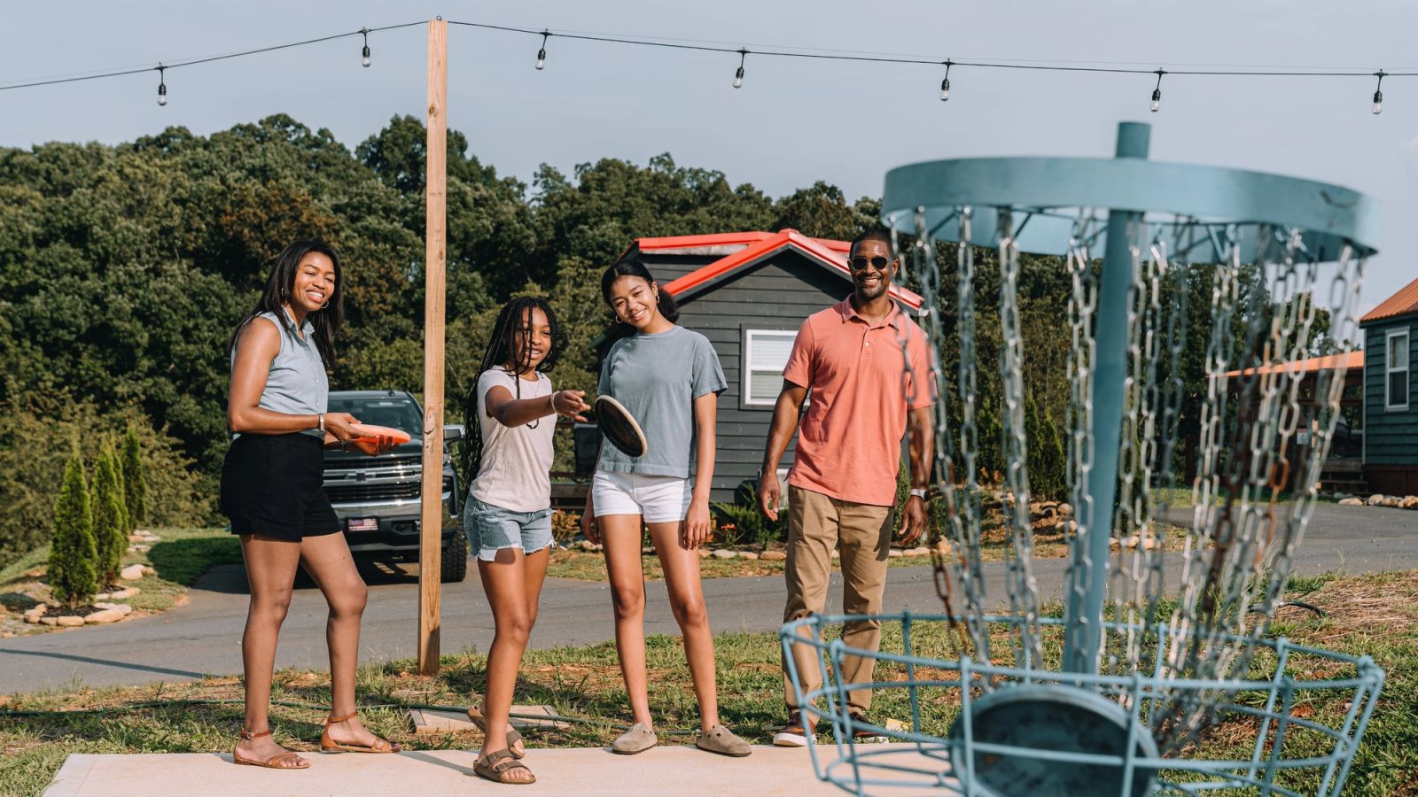 Four friends gathered around the disc golf basket beside the road at Sojourn Lodging