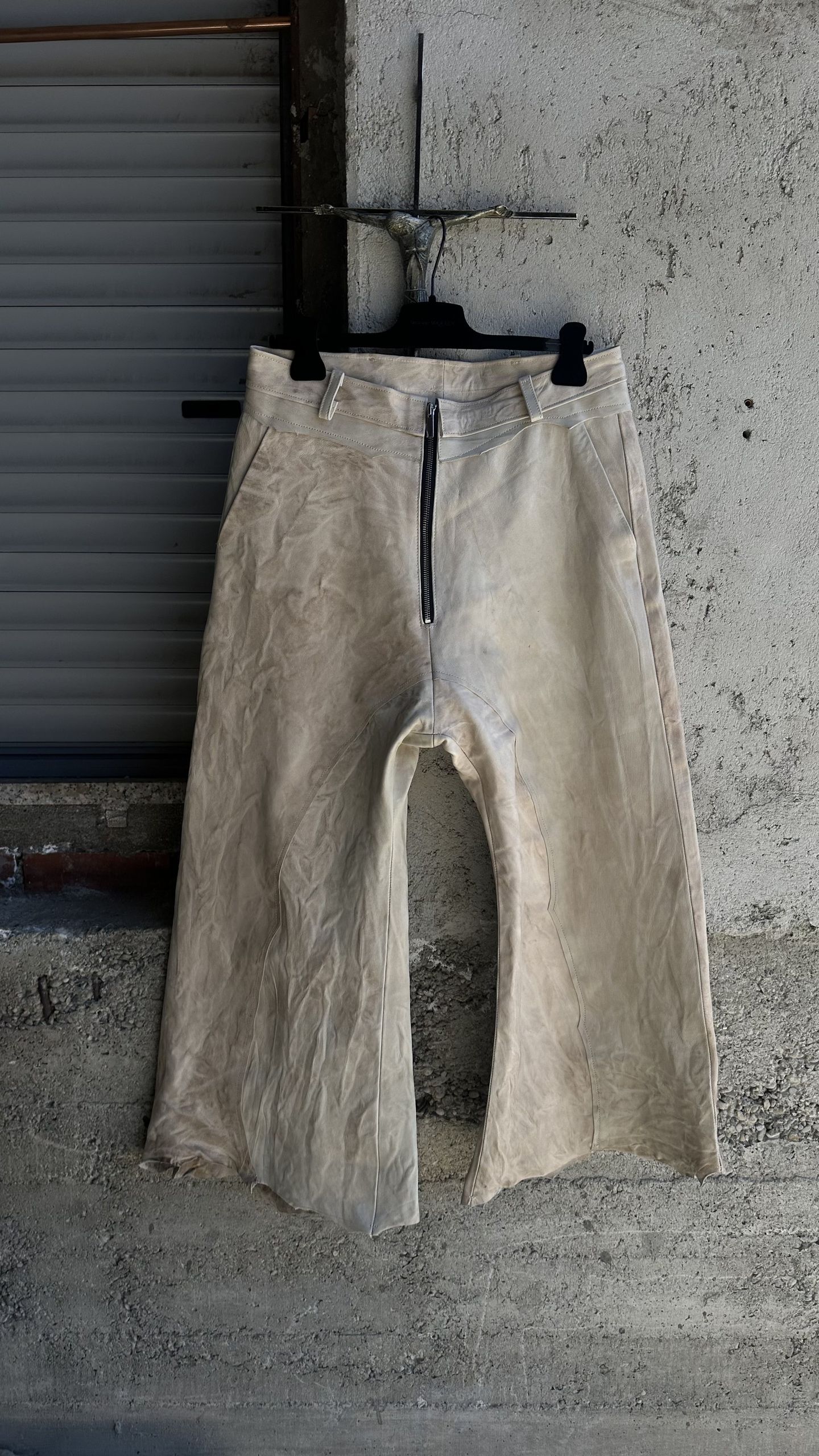 Pantaloni ampi in tela chiara, visti di fronte, appesi a una gruccia contro un muro di cemento e una serranda metallica.