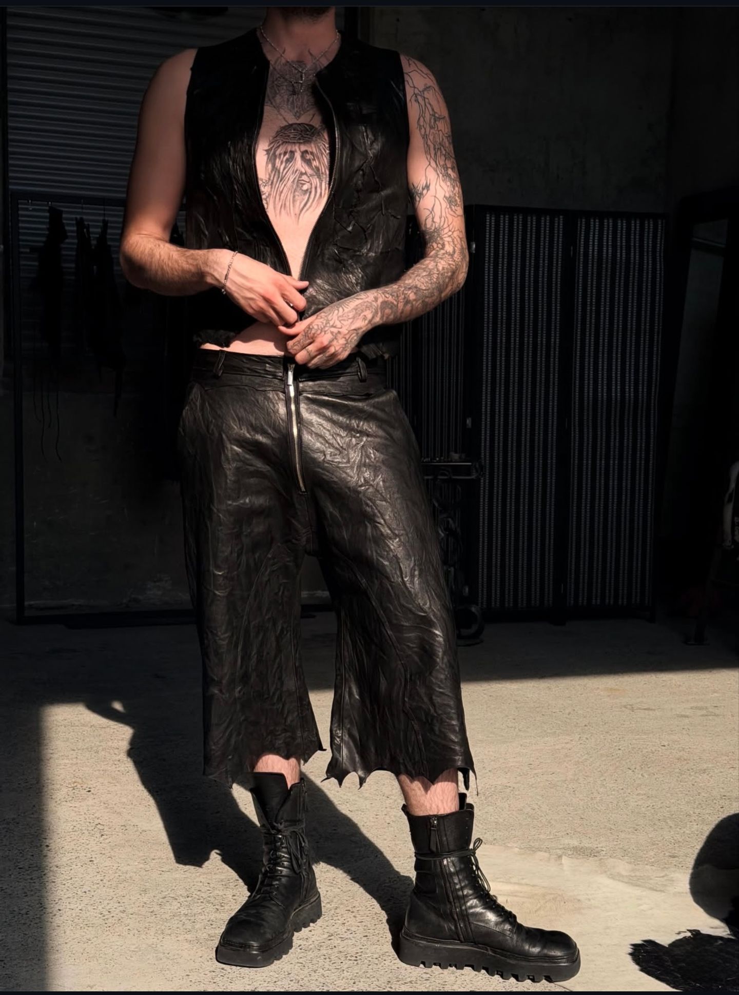 Uomo con gilet in pelle nera e pantaloni corti in pelle, al sole sul cemento.