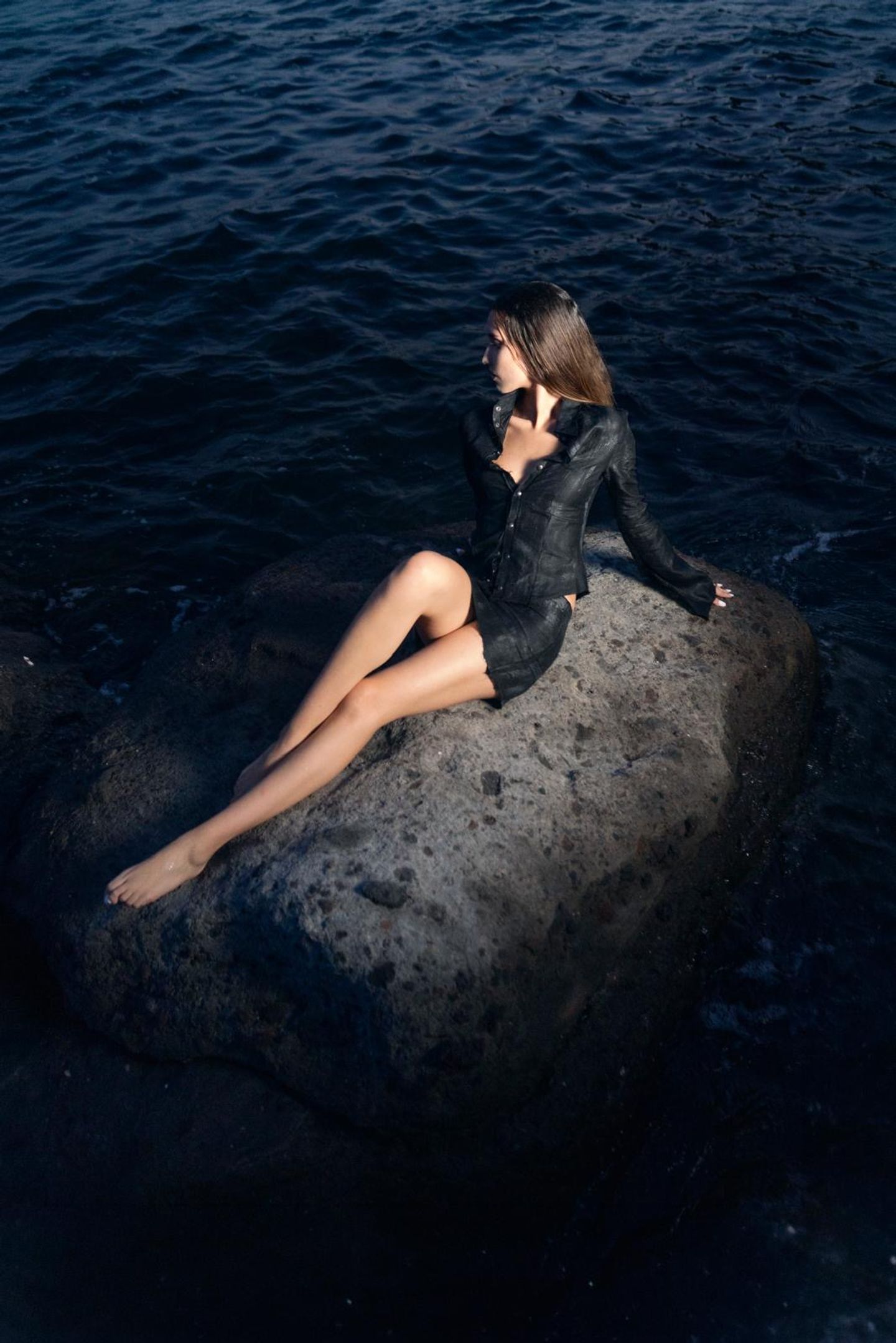 Modella distesa su uno scoglio in giacca di pelle scura, il mare aperto intorno.