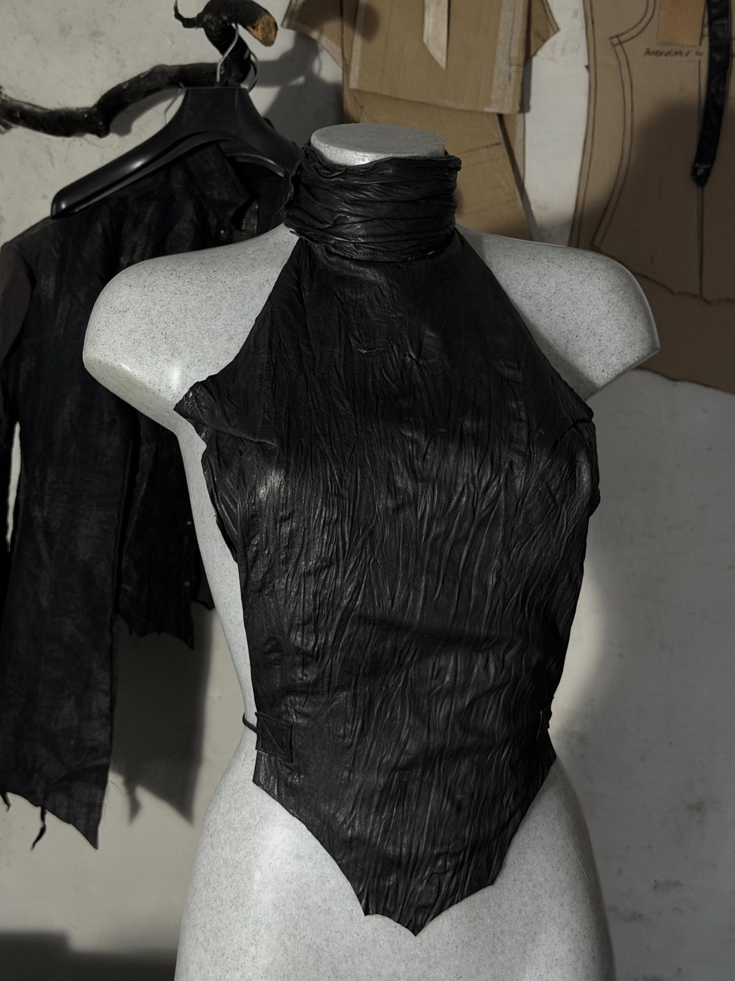 Top smanicato in pelle nera increspata con collo alto arricciato, su un manichino chiaro; a sinistra una camicia scura su una gruccia e sul muro dietro i cartamodelli.