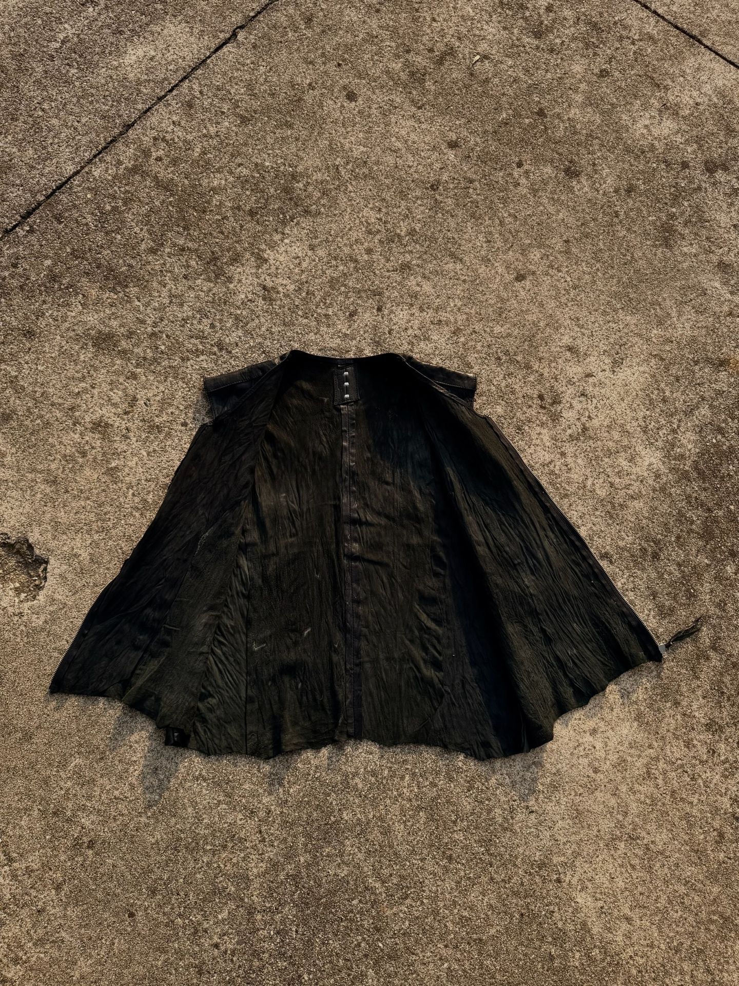 Capo in pelle nera aperto e disteso sul cemento, a forma di mantella.