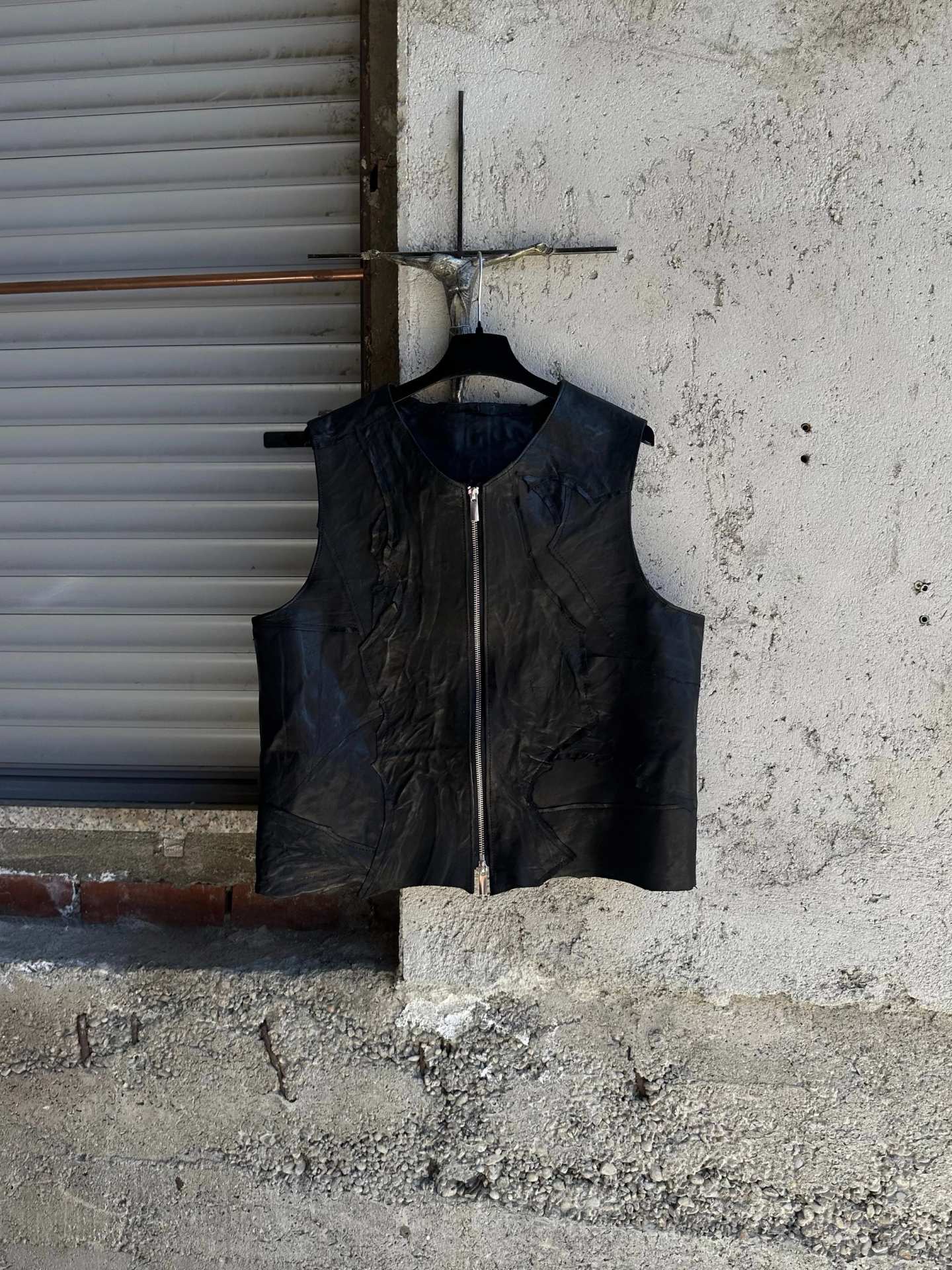 Gilet in pelle nera con zip centrale, appeso a una gruccia davanti a una serranda.