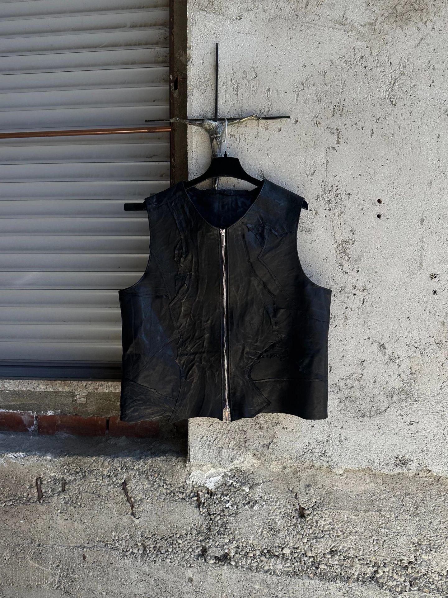 Gilet in pelle nera con zip centrale, appeso a una gruccia davanti a una serranda.