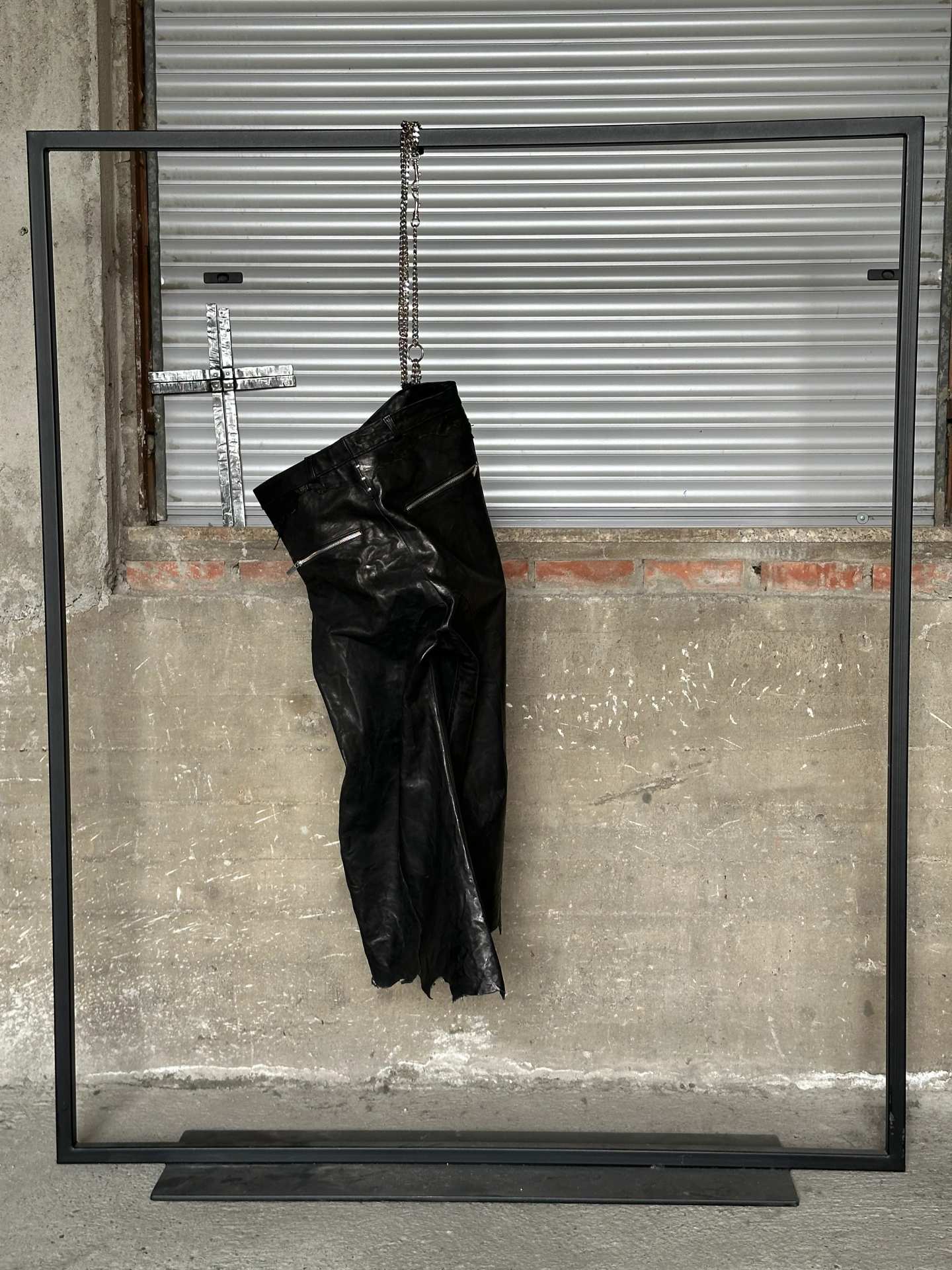 Pantaloni in pelle nera appesi a una parete di cemento, sotto una croce di metallo.