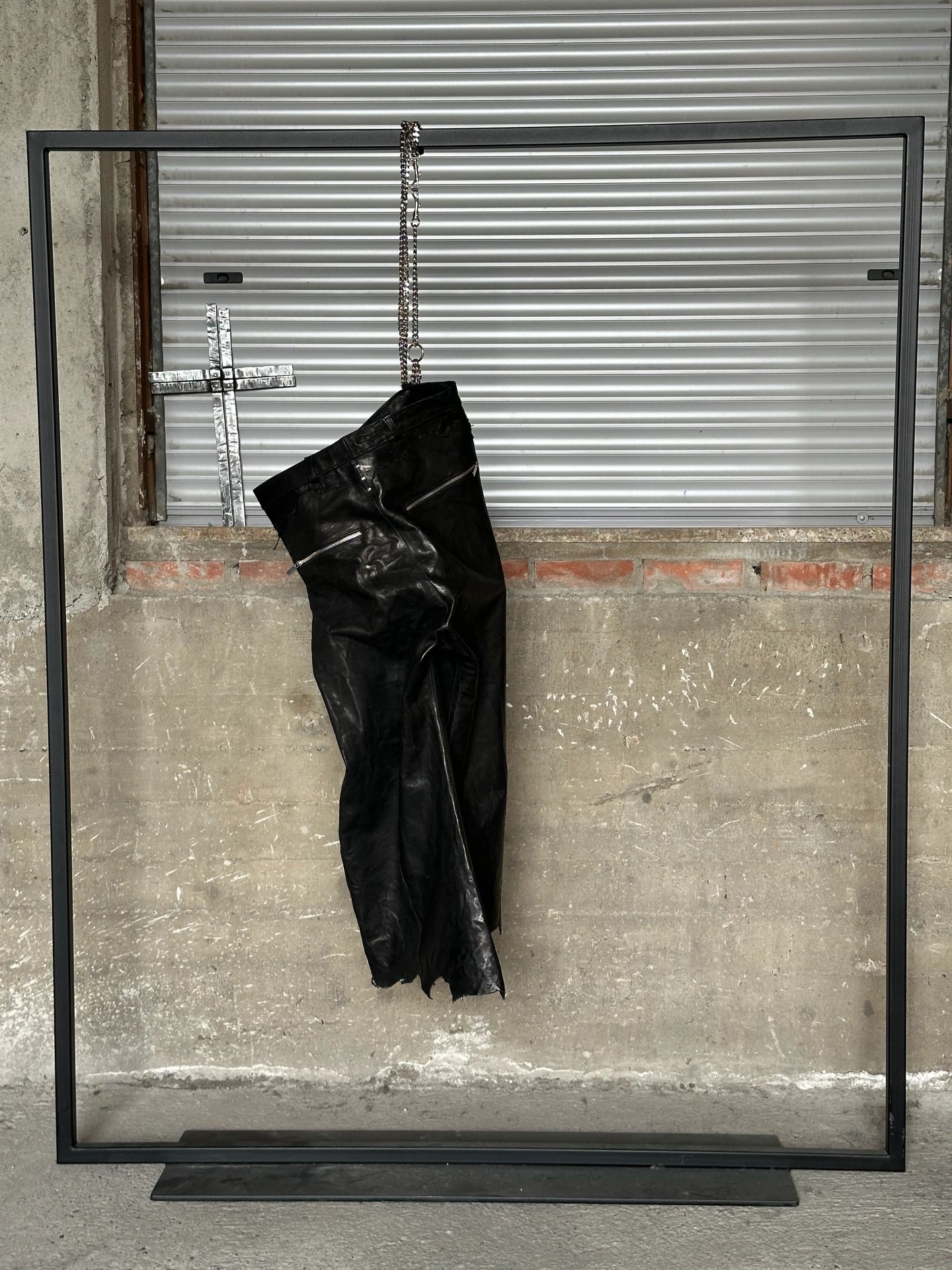 Pantaloni in pelle nera appesi a una parete di cemento, sotto una croce di metallo.
