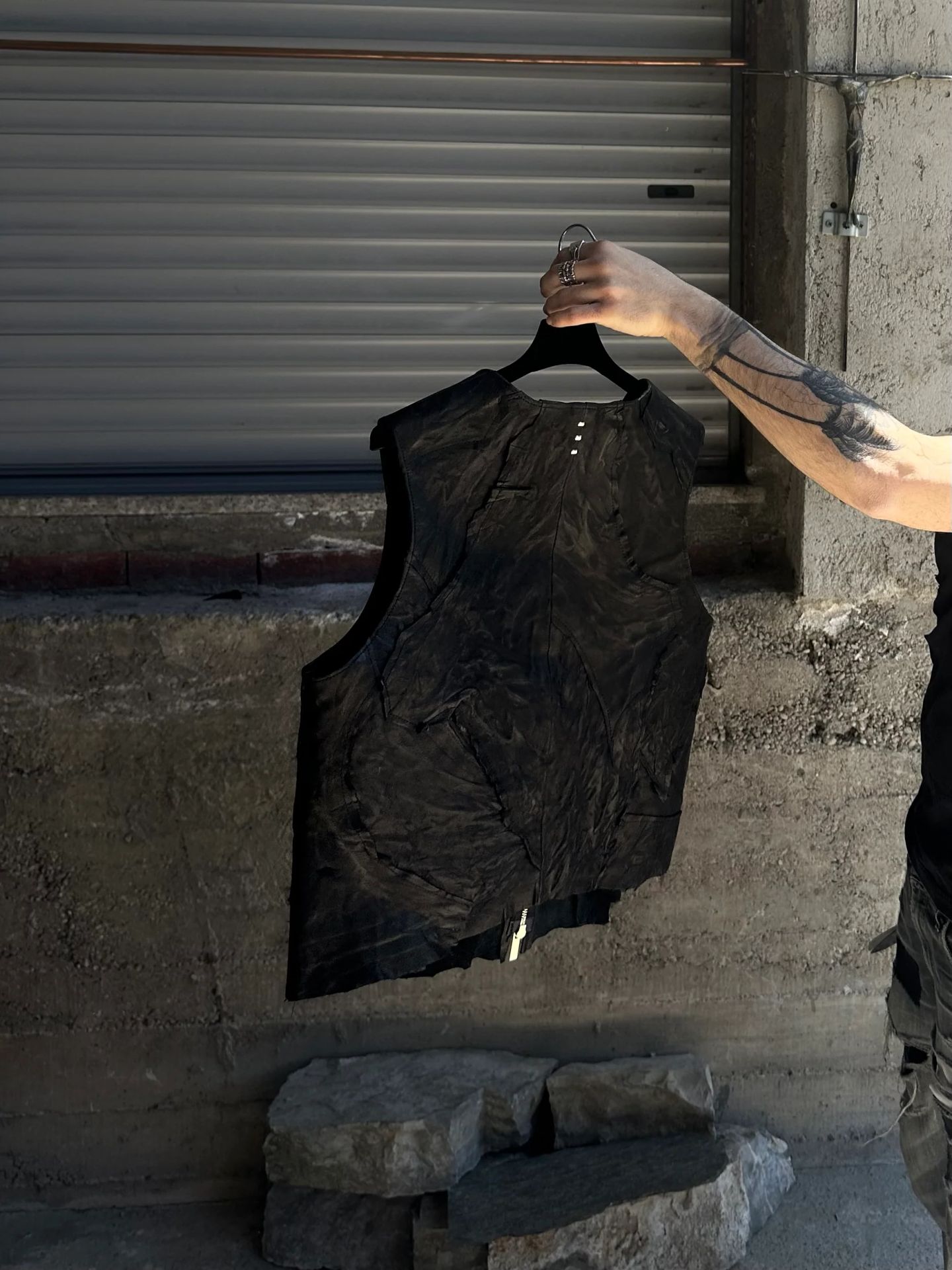 Gilet in pelle nera stropicciata, con bottoni automatici al collo, tenuto in mano davanti a una serranda.