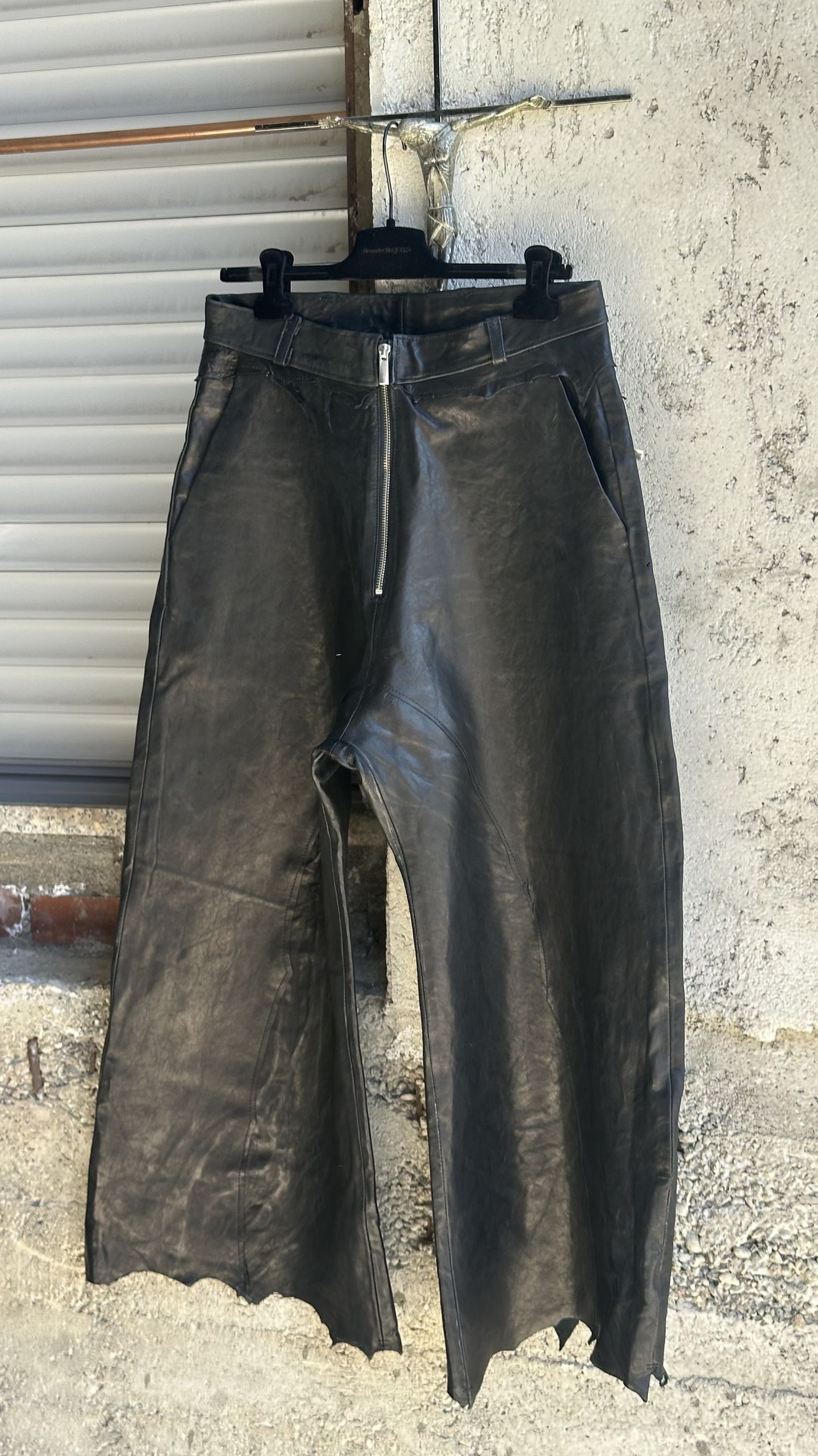 Pantaloni ampi in pelle nera, visti di fronte, con zip a vista e orlo grezzo, appesi contro un muro di cemento.