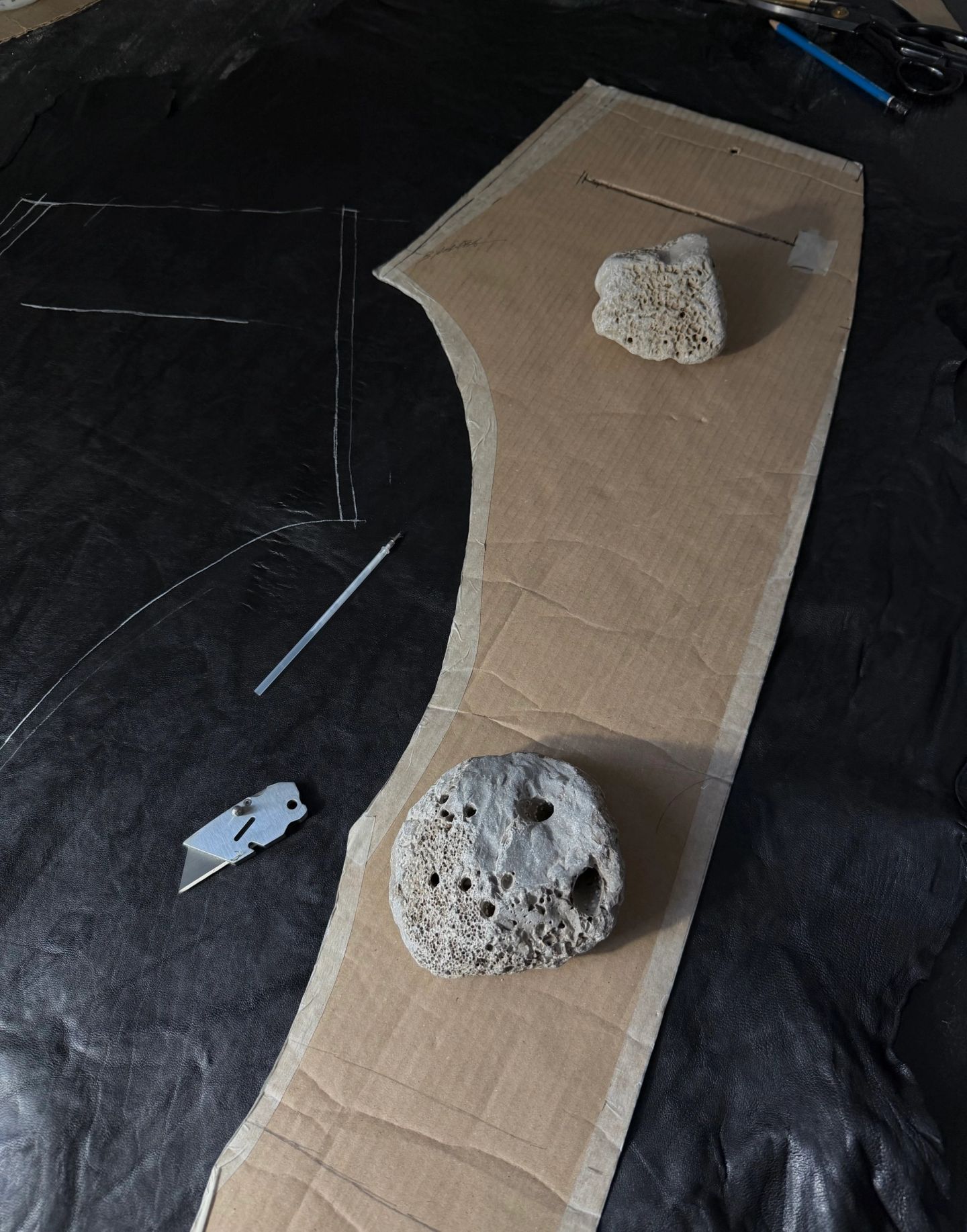 Un cartamodello di cartone appoggiato su una pelle nera, tenuto fermo da due pietre, con il taglio segnato a gesso.