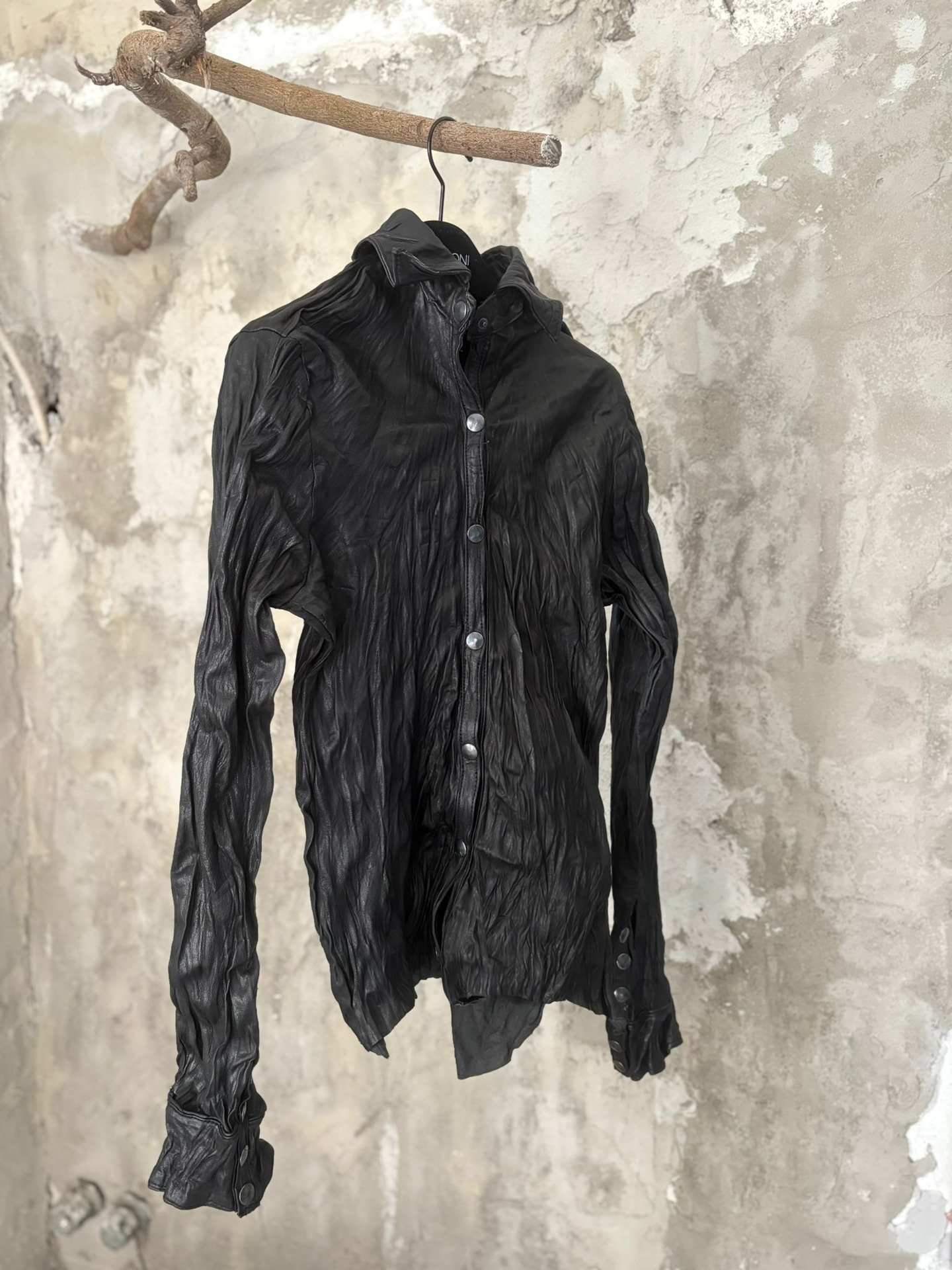 Camicia in pelle nera stropicciata, vista frontale, appesa a un muro di cemento chiaro.