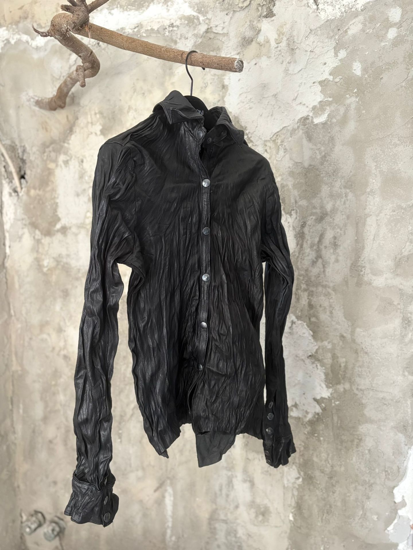 La camicia in pelle nera increspata, appesa a un ramo contro il muro di calce.