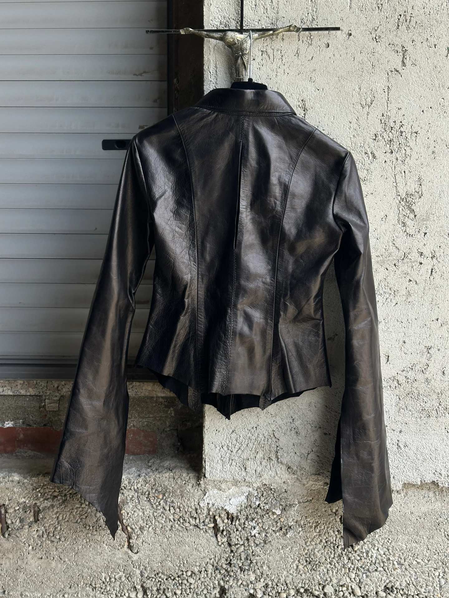 Camicia in pelle nera con orlo a punte, vista frontale, appesa davanti a una serranda metallica.