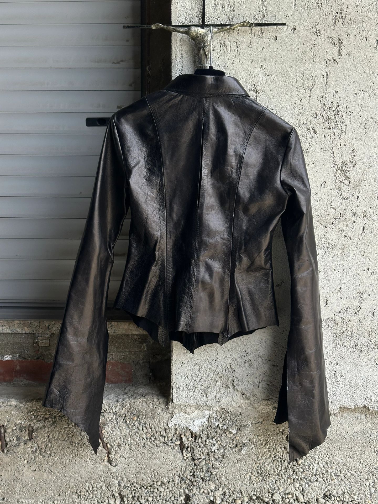 Camicia in pelle nera con orlo a punte, vista frontale, appesa davanti a una serranda metallica.