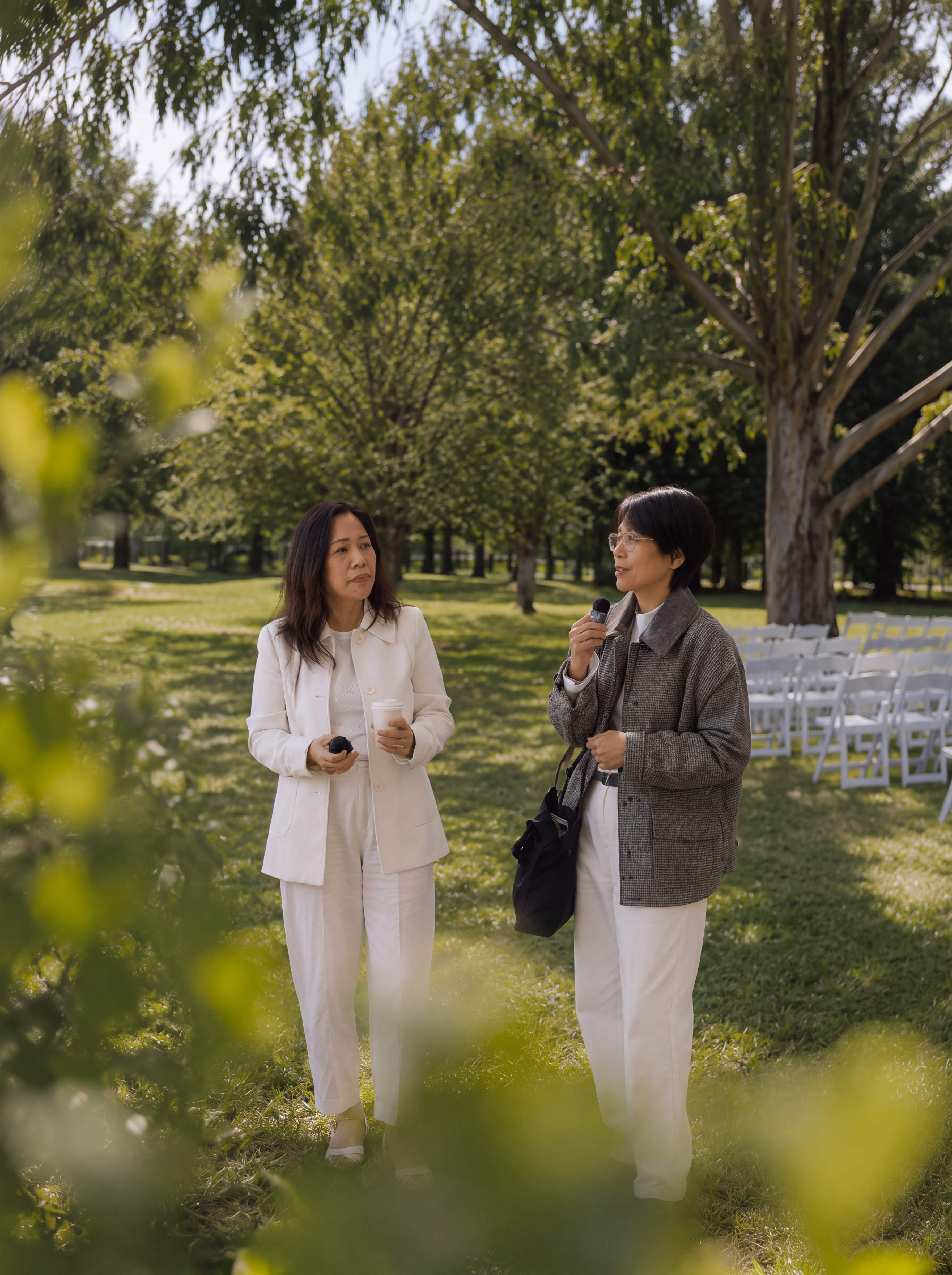Two women talking in a park while one holds a microphone, with white event chairs arranged in the background.