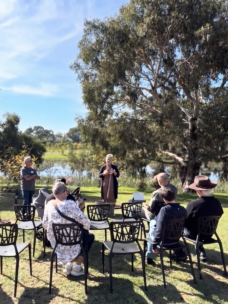 Open Day visitors gathered by a lake for a memorial tree ceremony under gum trees.