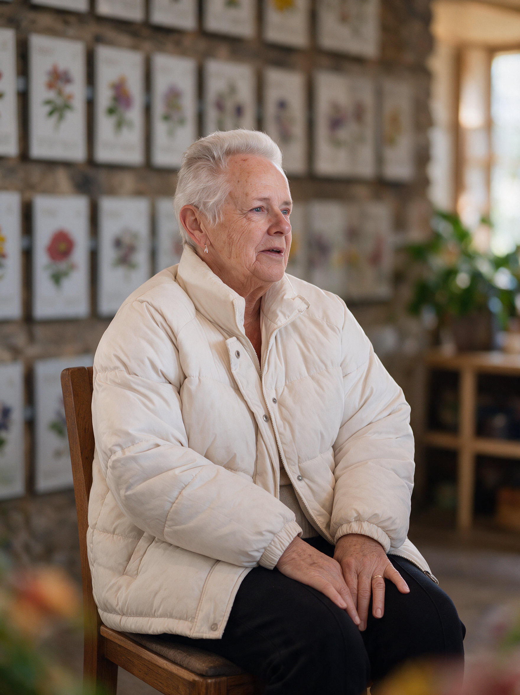 Elderly woman in a white puffer jacket seated in a wooden chair indoors.