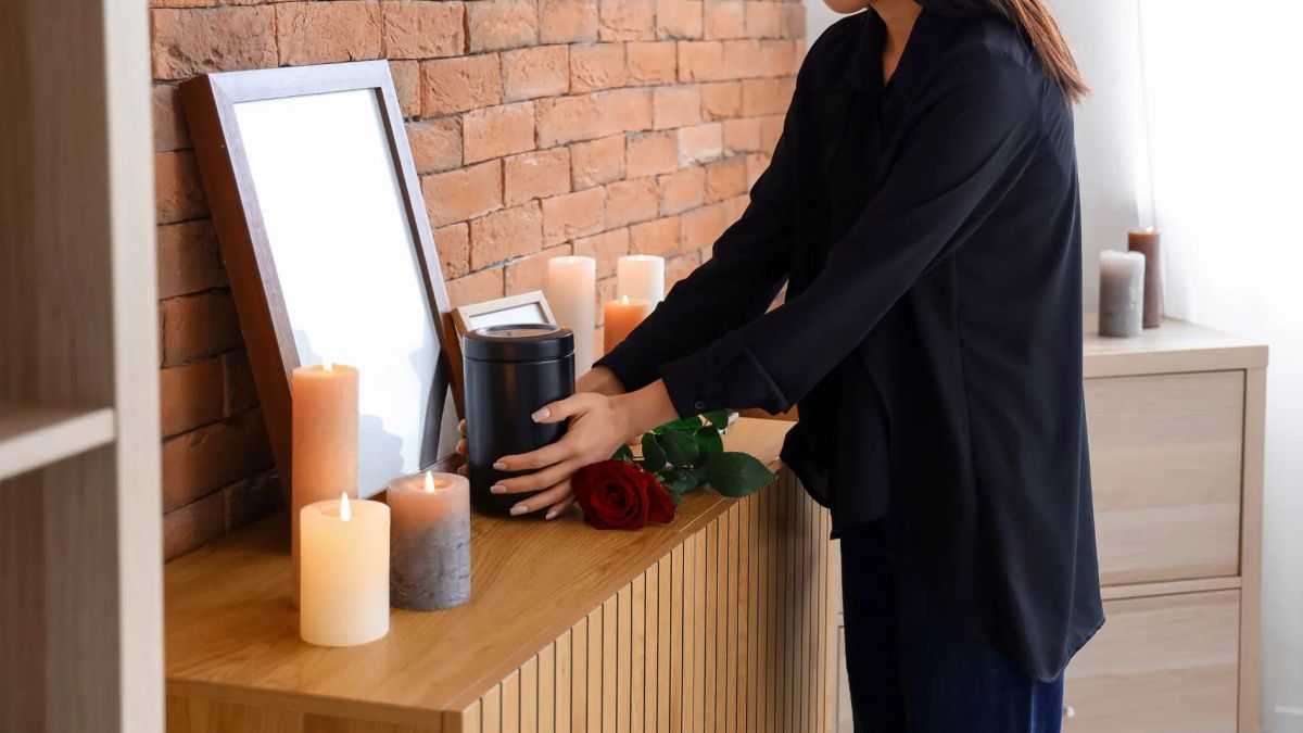 Woman placing a cremation urn on a home memorial shelf with candles, roses and a photo frame