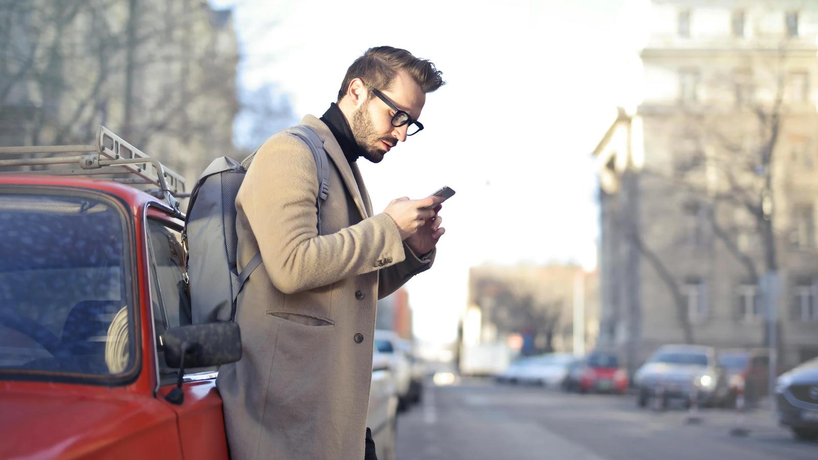 Мужчина в бежевом пальто держит телефон, опираясь на красный автомобиль