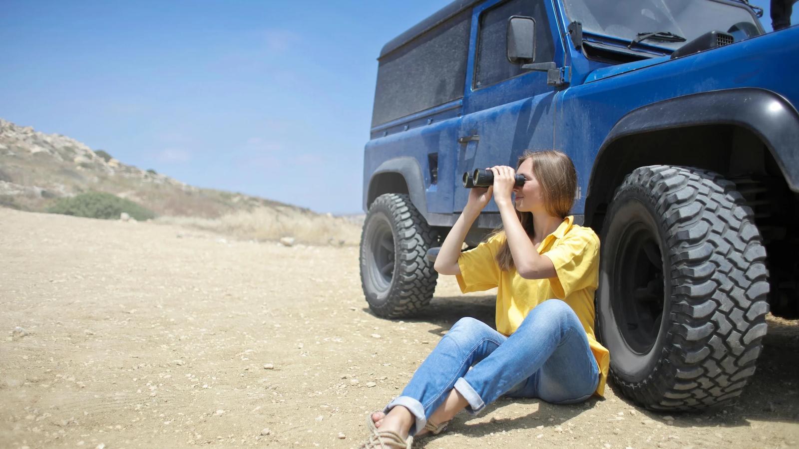 Женщина сидит на земле рядом с джипом, используя бинокль