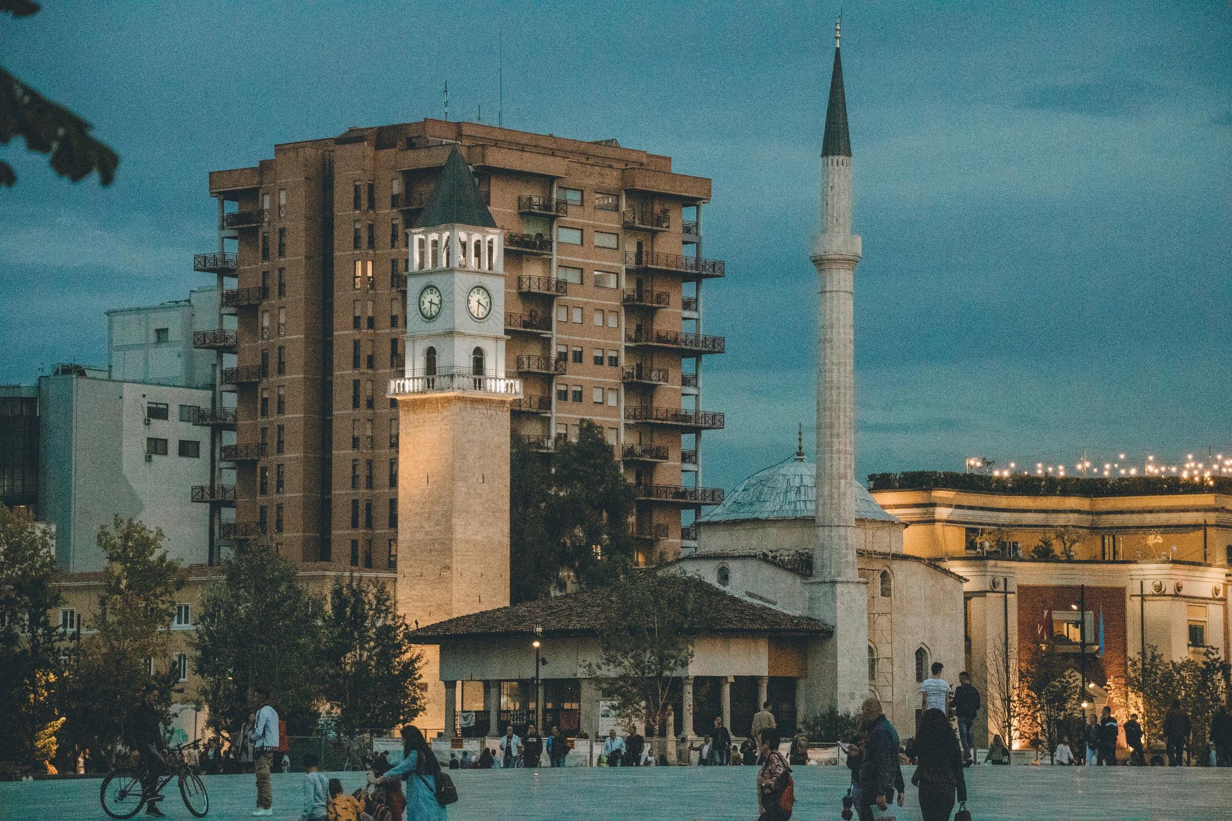 a group of people walking around a plaza with a clock tower in the background