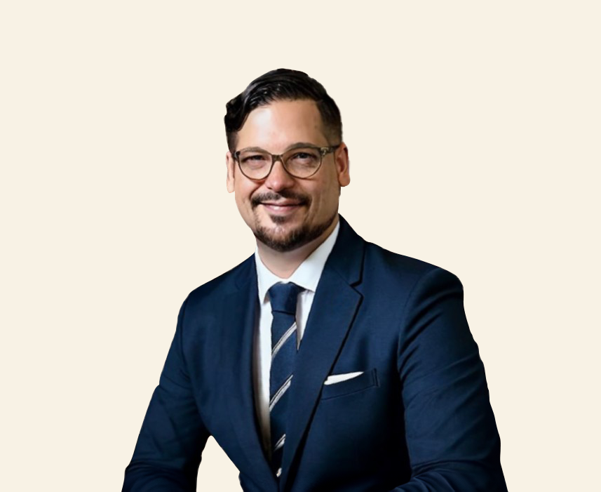 Photographic portrait of a dark-haired man in a navy blue suit, glasses and tie, smiling against a light background framed by a dark blue circle.
