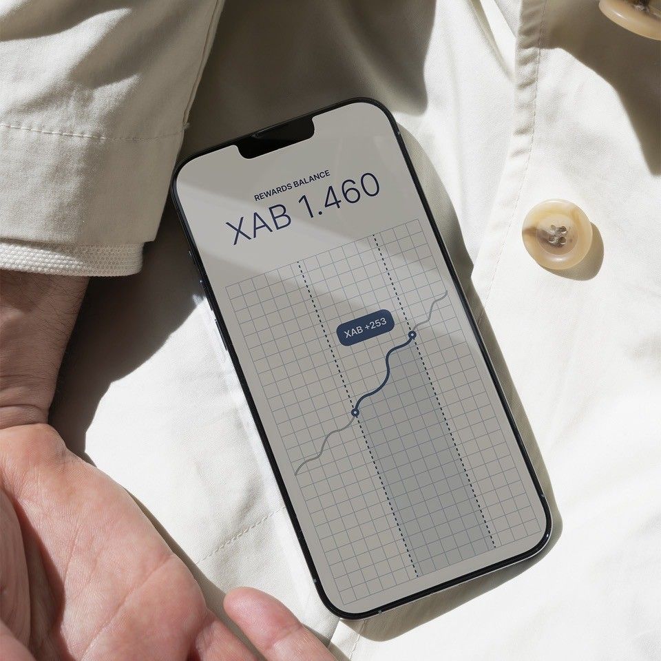 Close-up photo of a phone atop a cream-coloured garment. The screen shows a rewards balance of XAB 1460 with a graph showing a recent gain of XAB 253.