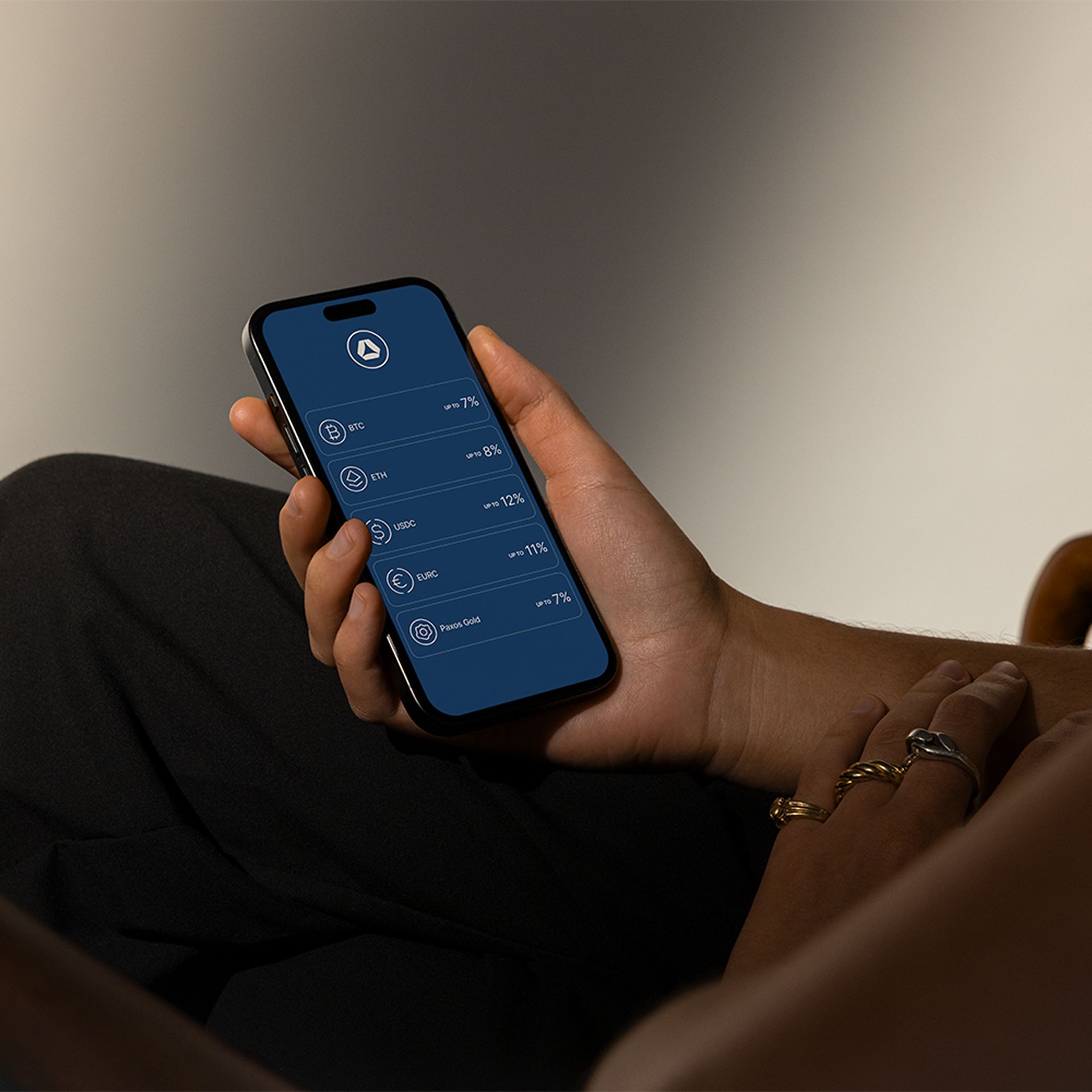 Photo of a person holding a smartphone displaying crypto assets like BTC, ETH and USDC with yield rates up to 12% on a dark blue app interface.