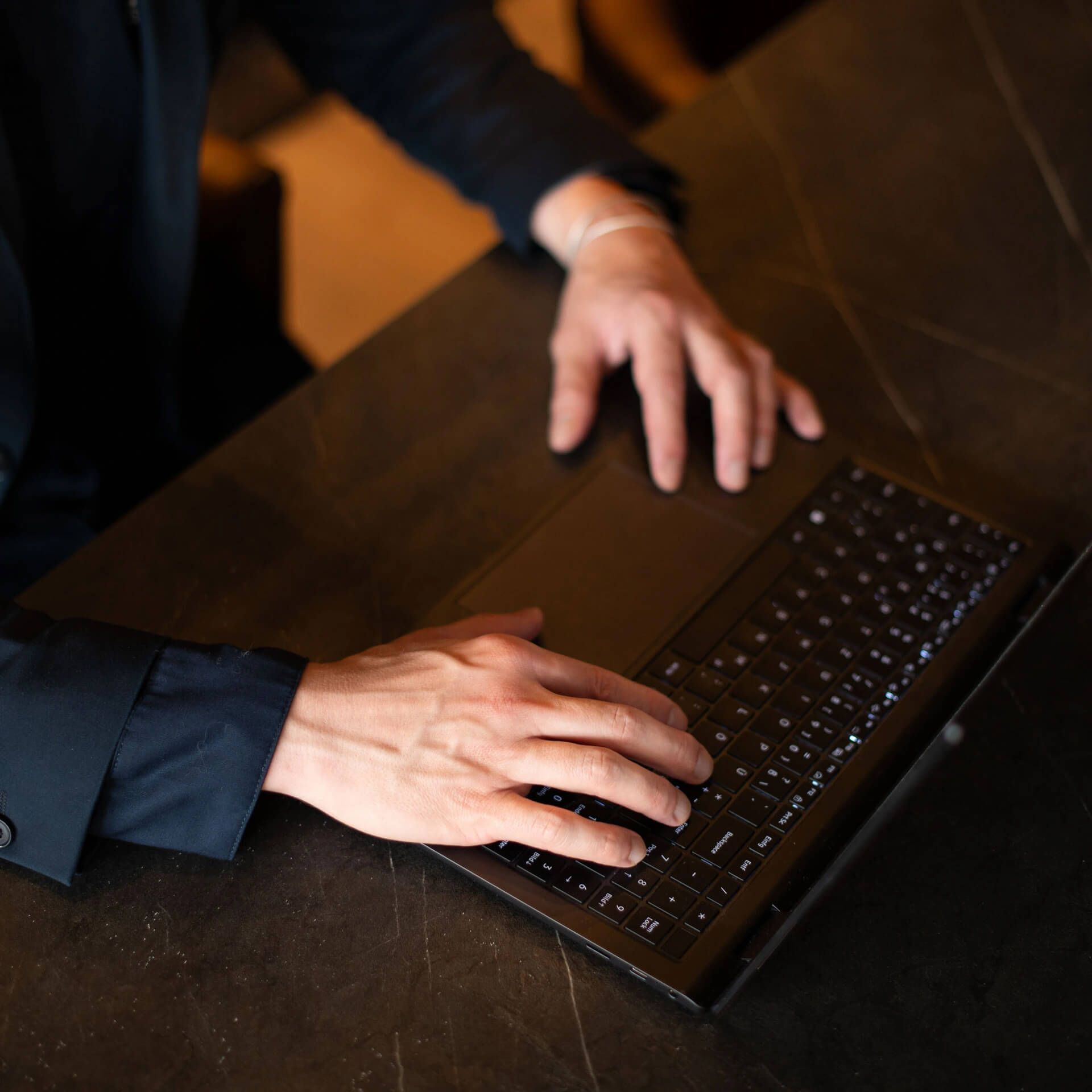 Close-up photo of a person's hands typing on a laptop keyboard on a dark wooden surface in warm lighting.