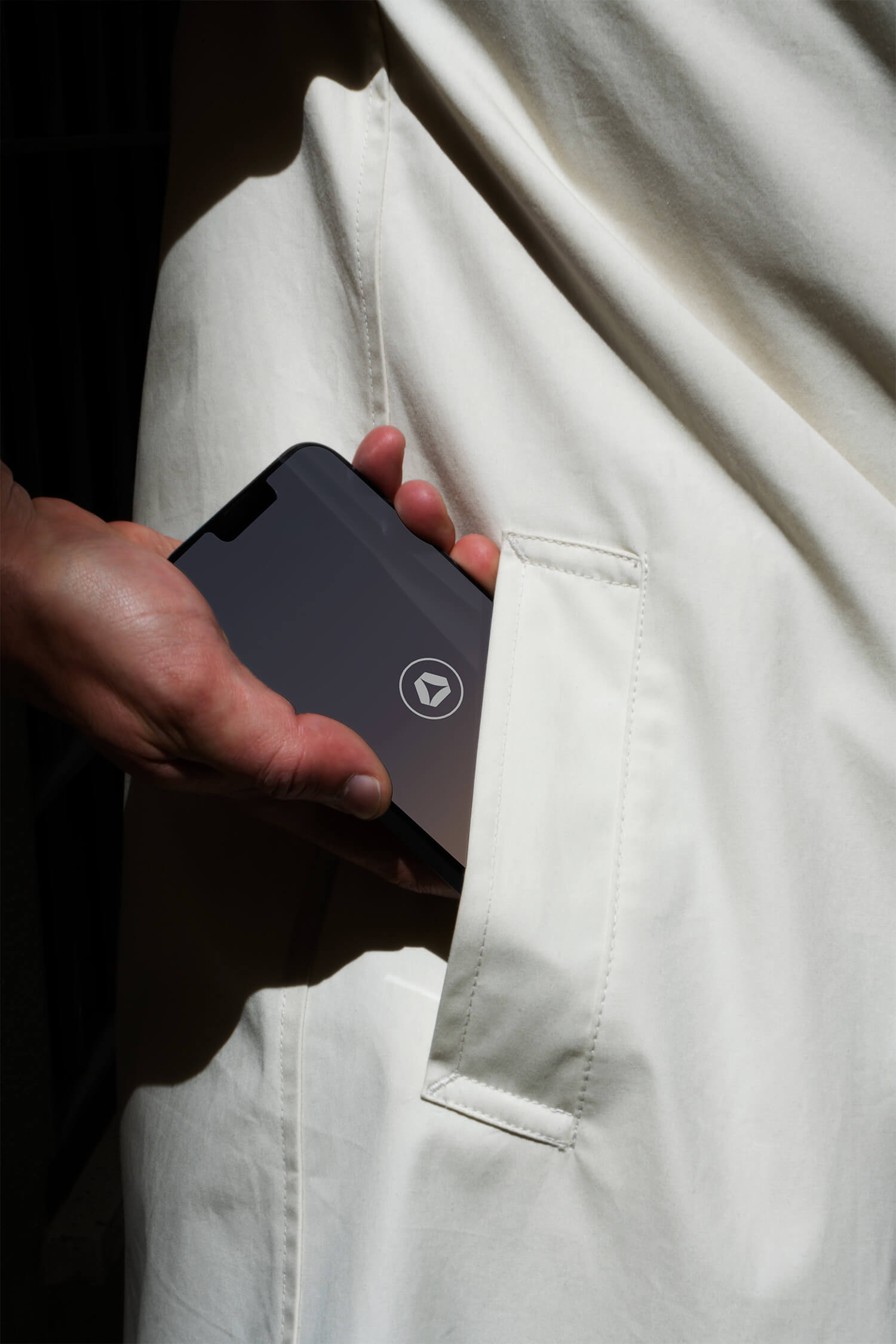 Close-up photo of a hand sliding a smartphone into the pocket of a cream-coloured jacket. The phone screen has the Abloxx logo at the center. 
