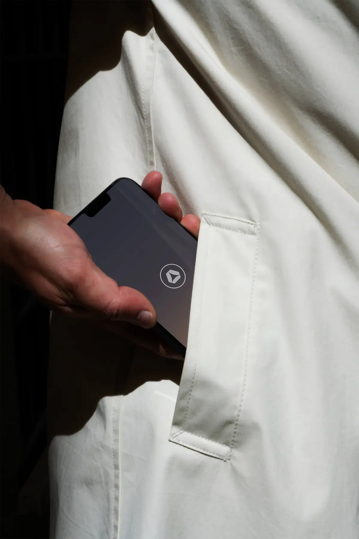 Close-up photo of a hand sliding a smartphone into the pocket of a cream-coloured jacket. The phone screen has the Abloxx logo at the center.