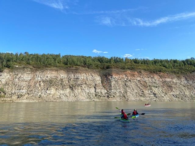 Two kayakers paddle near rocky cliff on a scenic river.