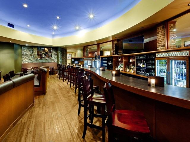 Modern hotel bar with wooden counters, seating, and illuminated drink displays.