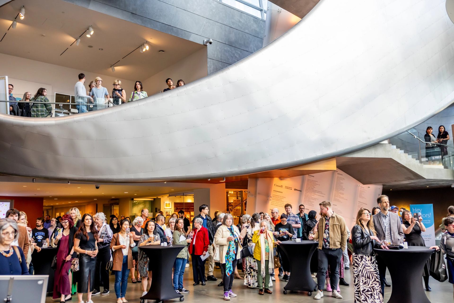 Crowd gathers around cocktail tables beneath a curved ramp in a modern atrium.