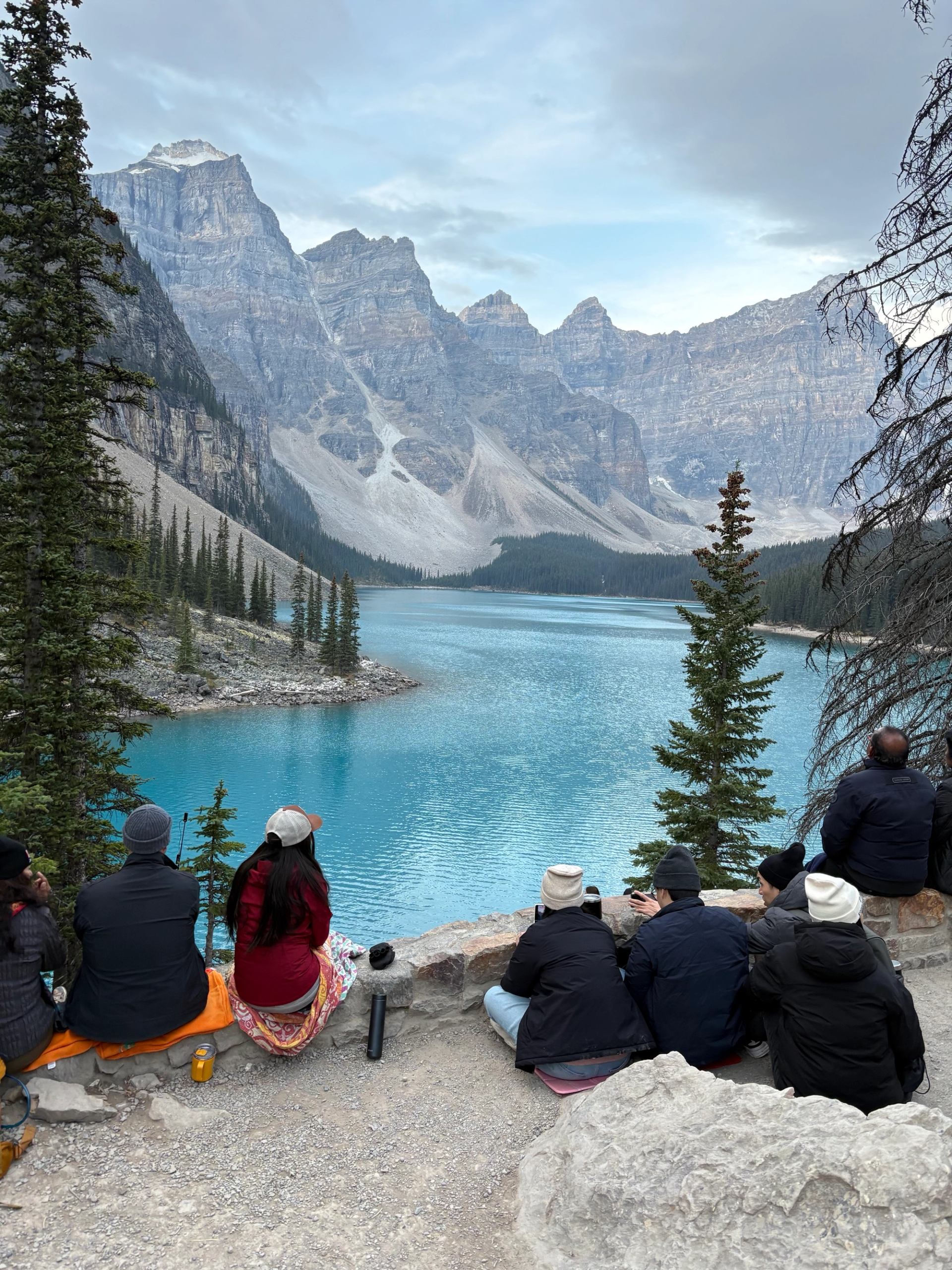 Group of people sitting by turquoise mountain lake, facing rocky peaks and evergreen trees.
