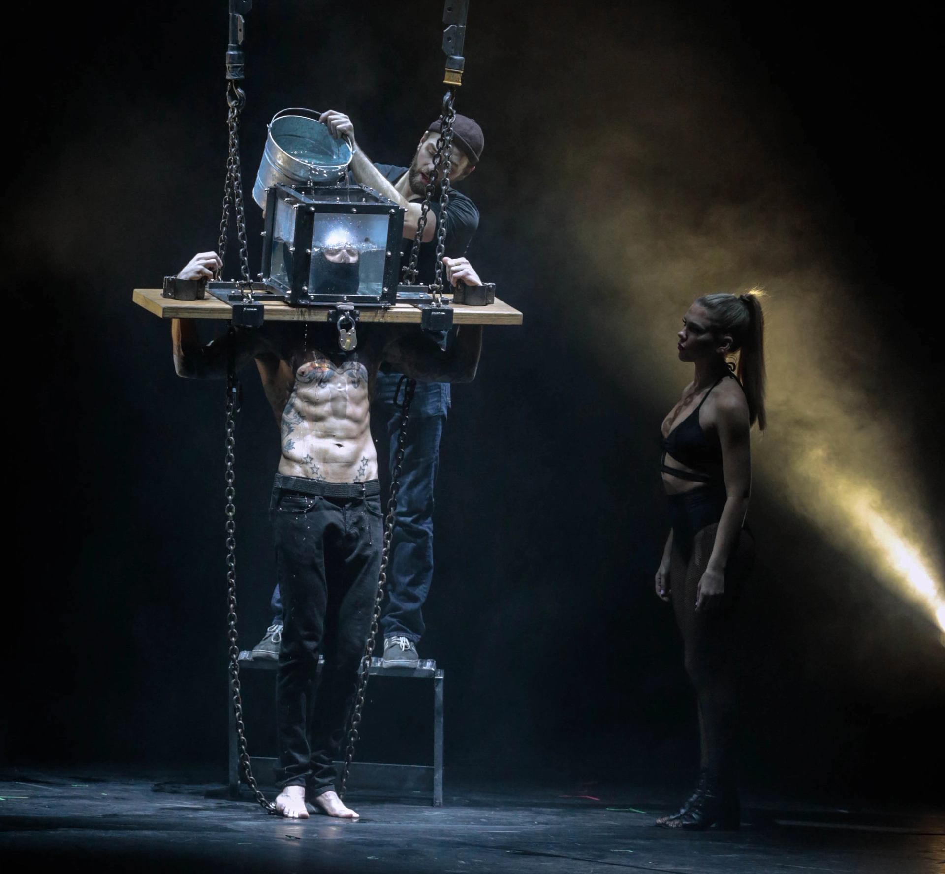 Stage escape act with performer in chained water tank apparatus and two assistants watching.