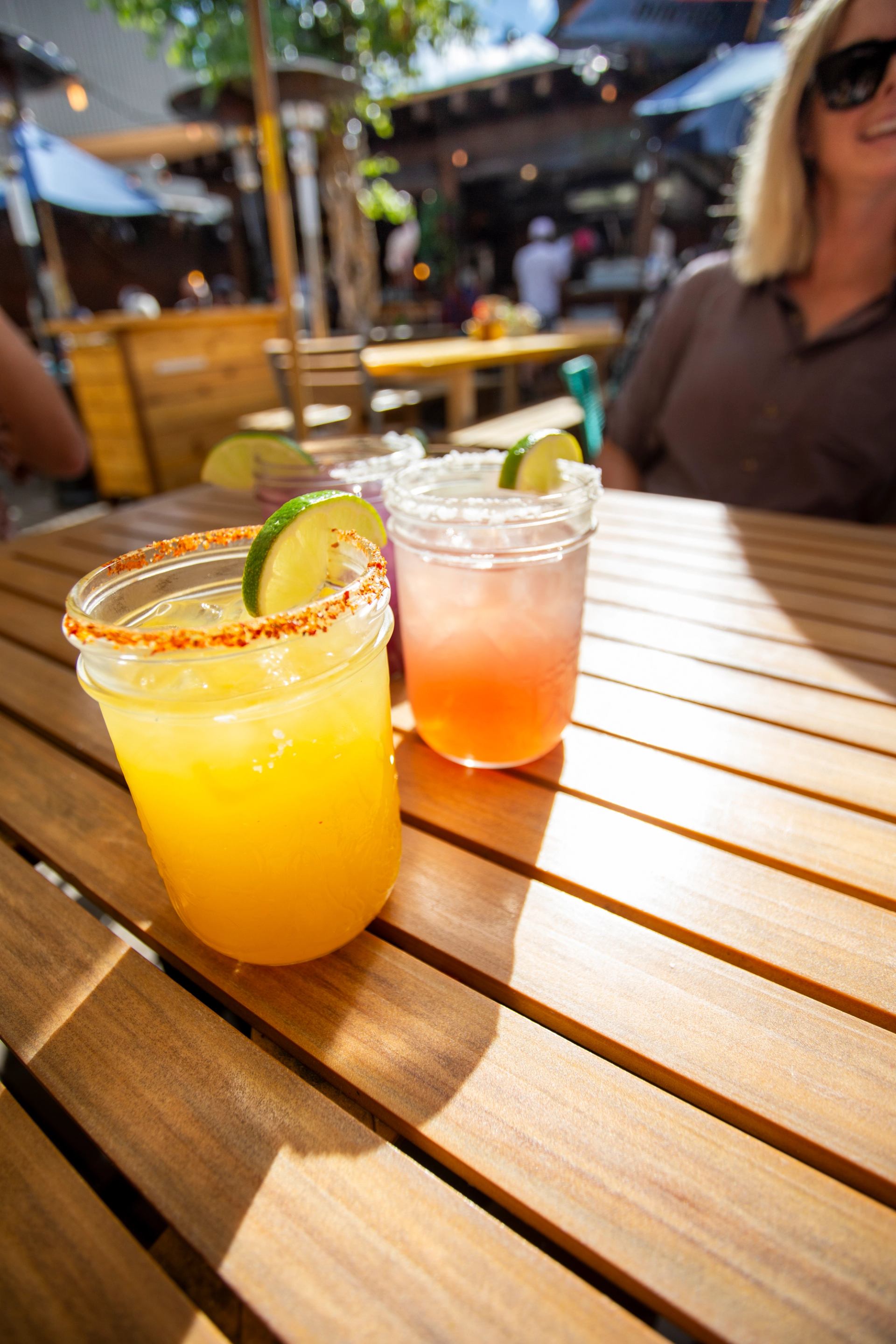 Two vibrant cocktails with lime and salt rims sit on a wooden outdoor table, with a woman in the blurry background.