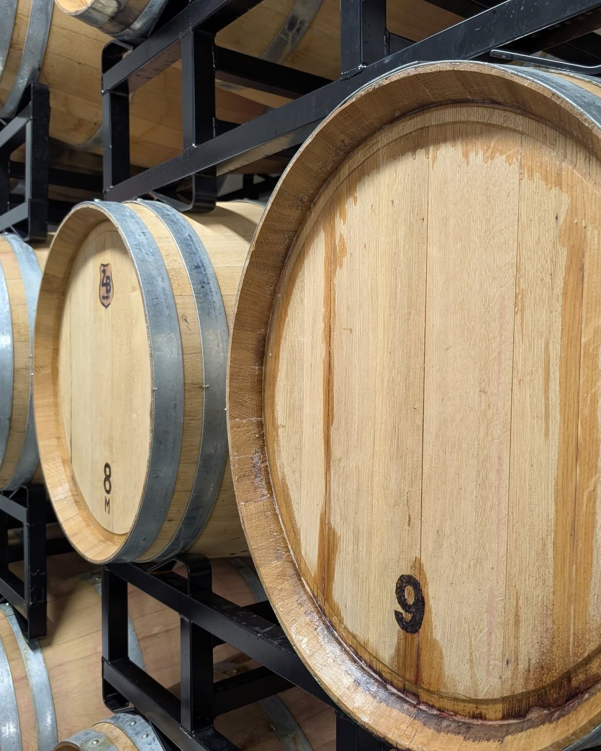 Wooden barrels with metal hoops stacked on black racks, front barrel marked 9.