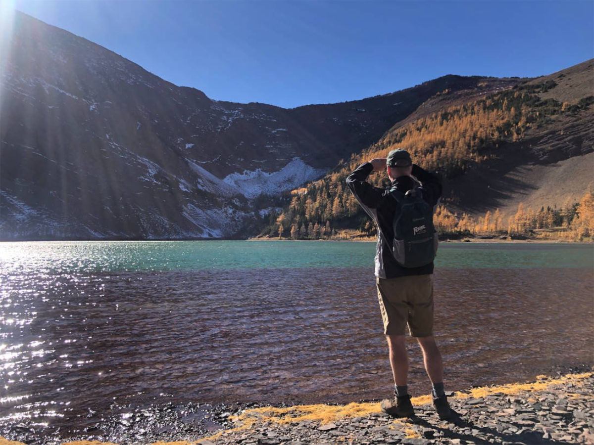 Person standing by clear alpine lake with mountains and autumn trees at Upper Rowe Lake.