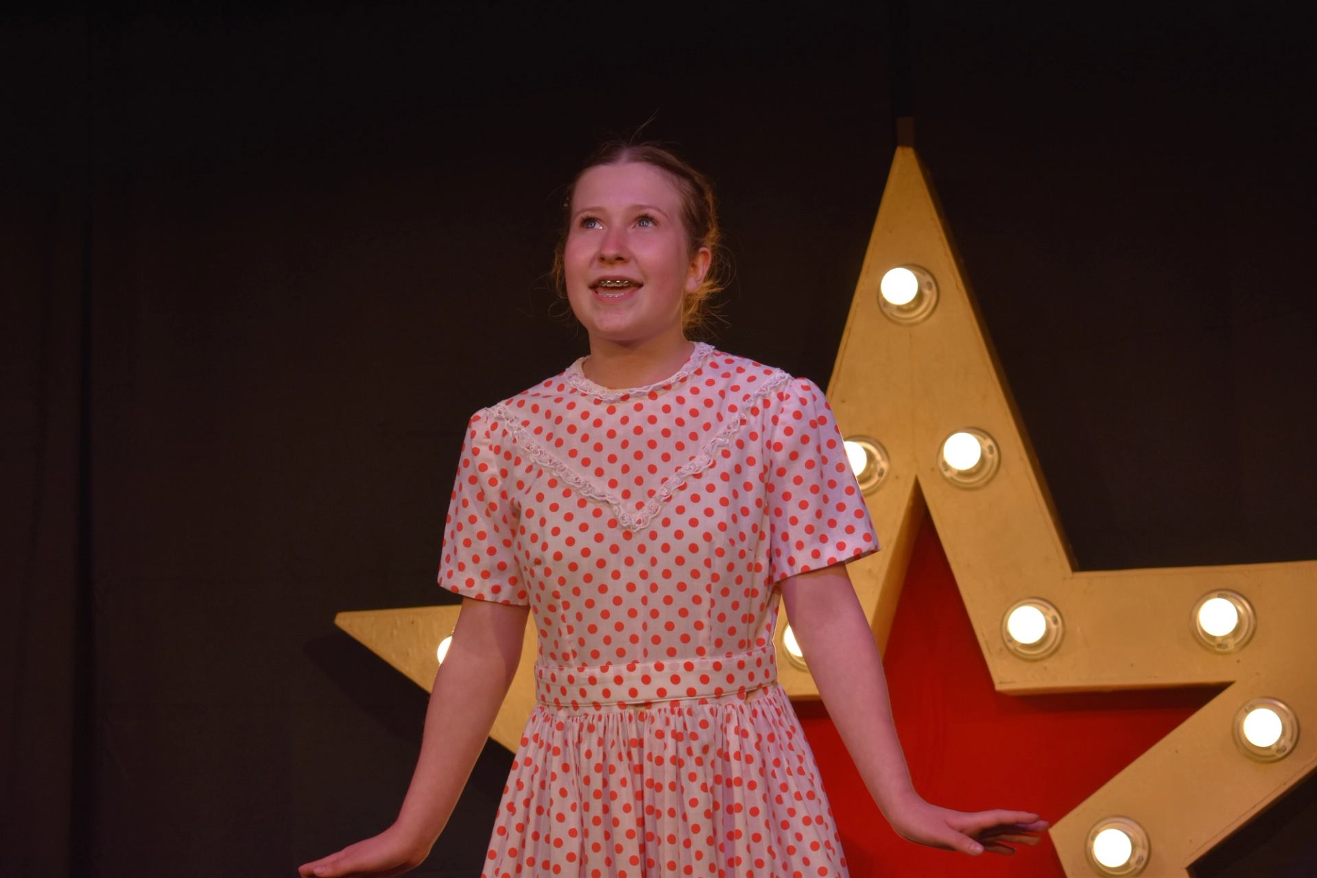 Performer in polka-dot dress stands before lit star on dark stage.