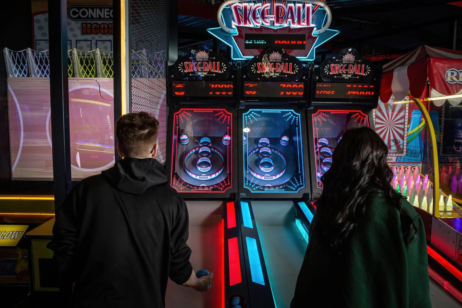 Two people playing skee-ball at an arcade with bright neon lights and colorful game machines.